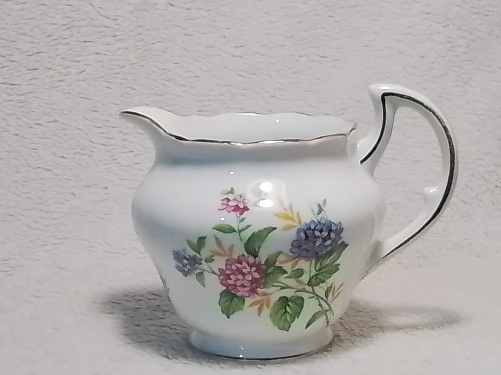 A white porcelain creamer with floral patterns of pink, purple, and green leaves, placed on a light-colored textured surface.