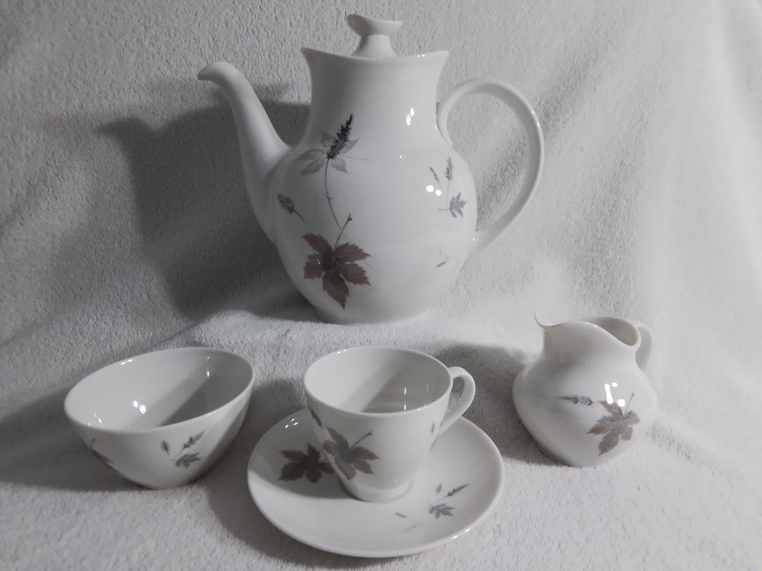 White ceramic tea set with brown leaf patterns, including a teapot, creamer, cup on saucer, and two bowls, displayed on a white textured surface.