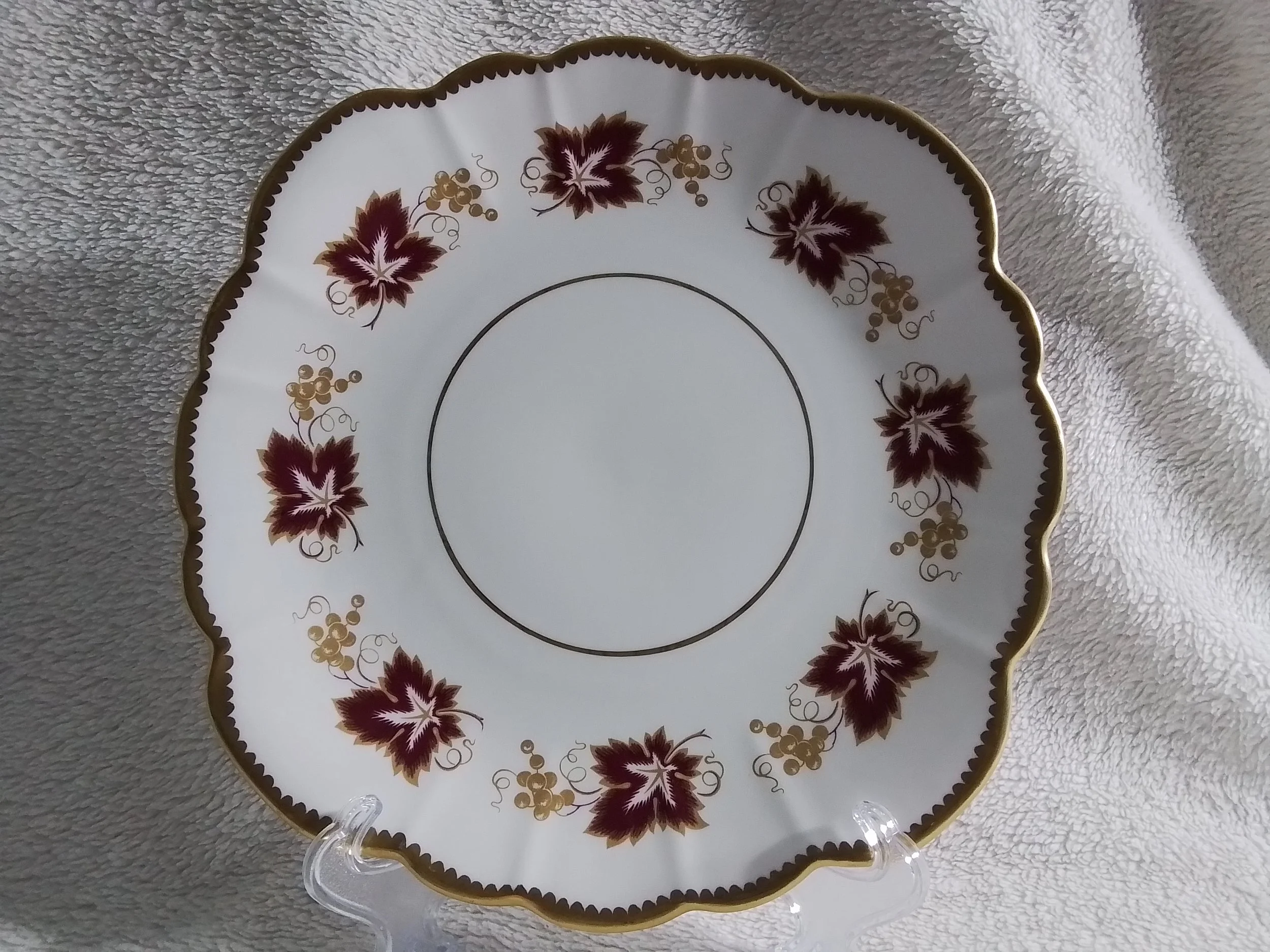 Decorative porcelain plate with a scalloped edge, gold trim, and a pattern of dark red leaves and gold grapes around the rim, displayed on a clear stand.