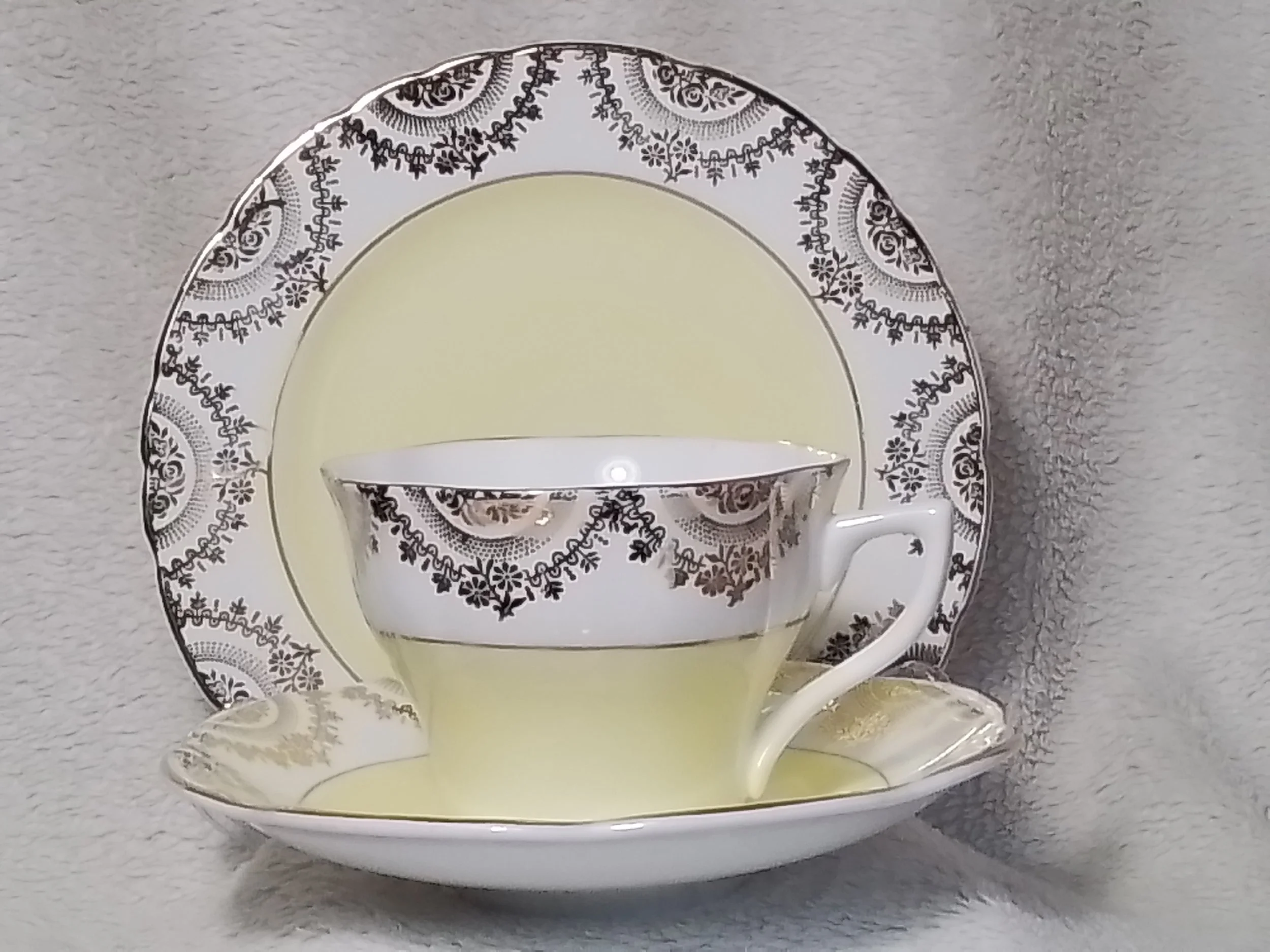 A fine china tea cup with a black floral pattern and gold accents, accompanied by a matching saucer and a large serving plate, all featuring a similar black floral design and a pale yellow band around the rim, set against a soft gray textured backgro