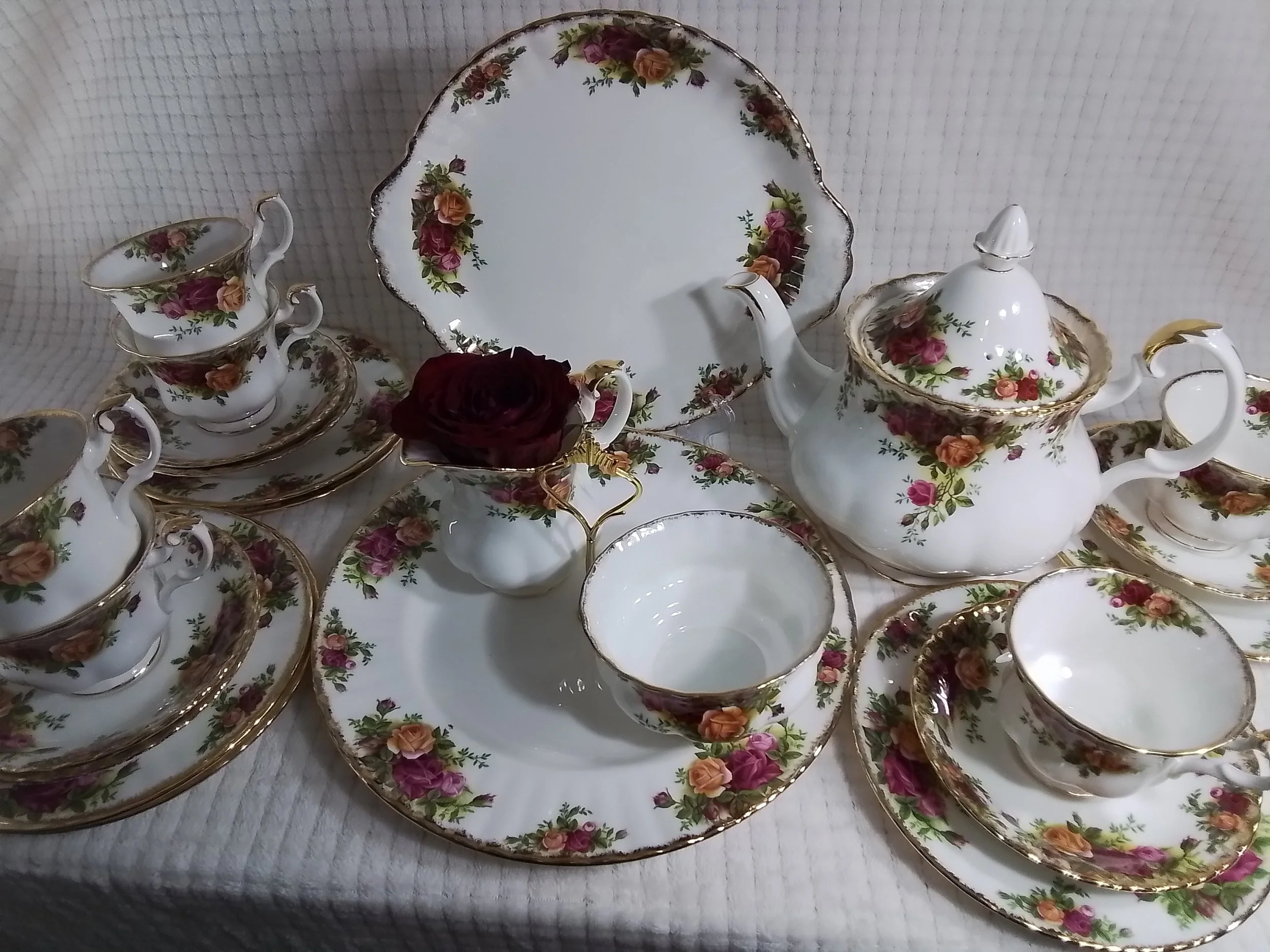 A collection of vintage floral-patterned china teacups, saucers, a teapot, and a cake plate on a textured white tablecloth with a dark red rose in one of the teacups.