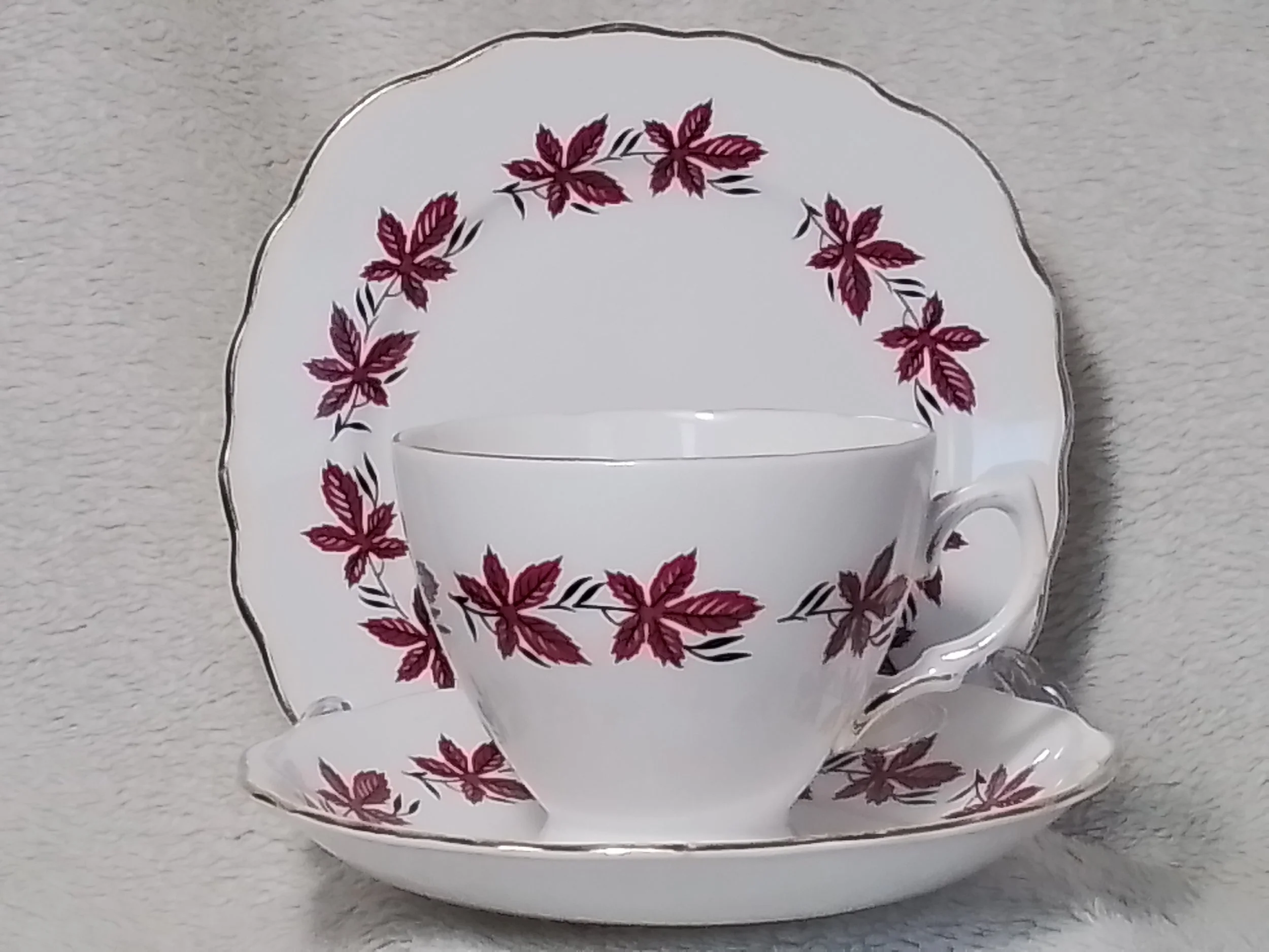A white china tea cup with a handle, placed on a matching saucer, both decorated with a pattern of red and black leaves. Behind them is a round plate with the same leaf pattern, set against a textured light-colored background.