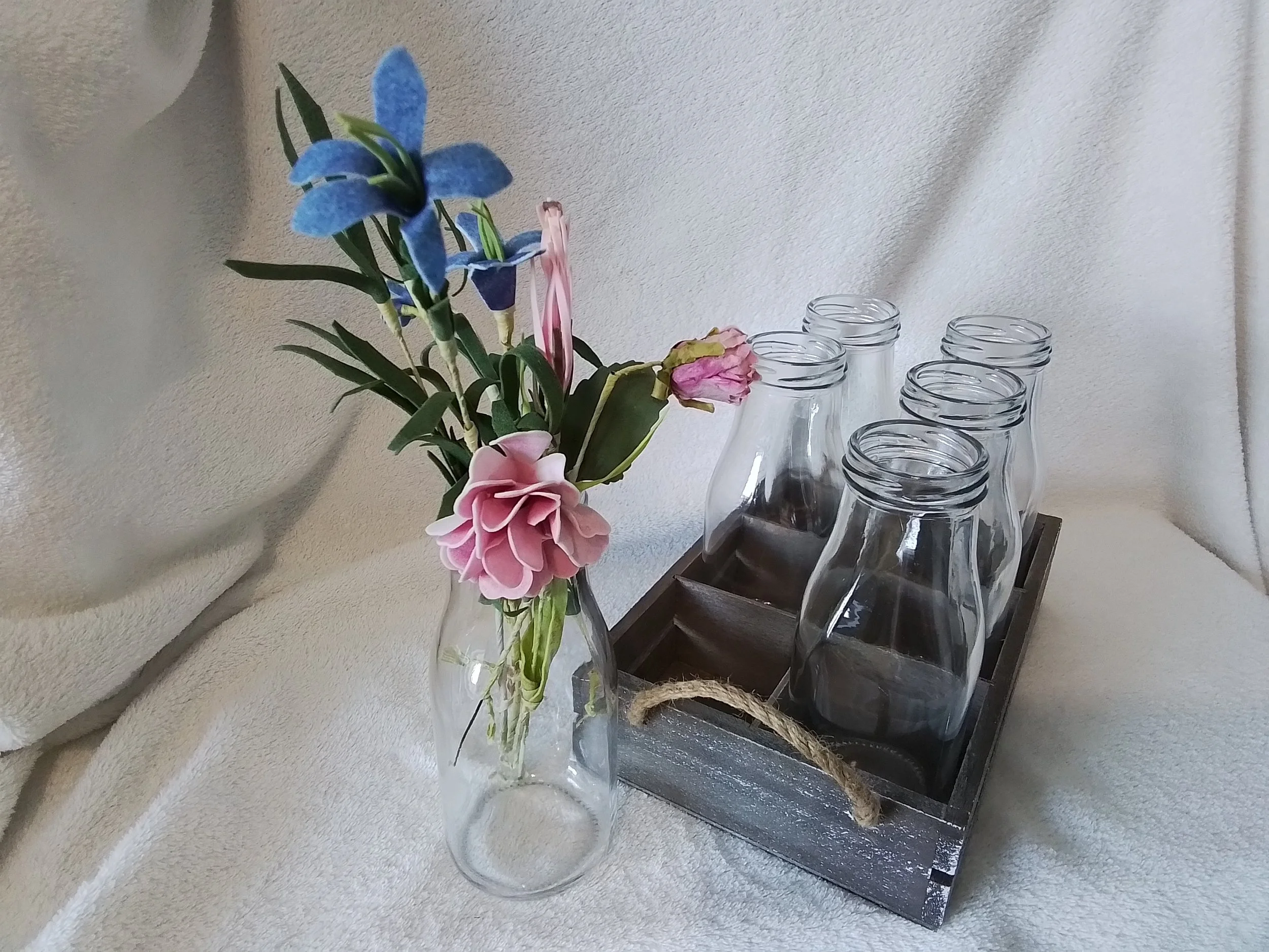 Glass vase with pink and blue artificial flowers next to a wooden tray holding empty glass bottles
