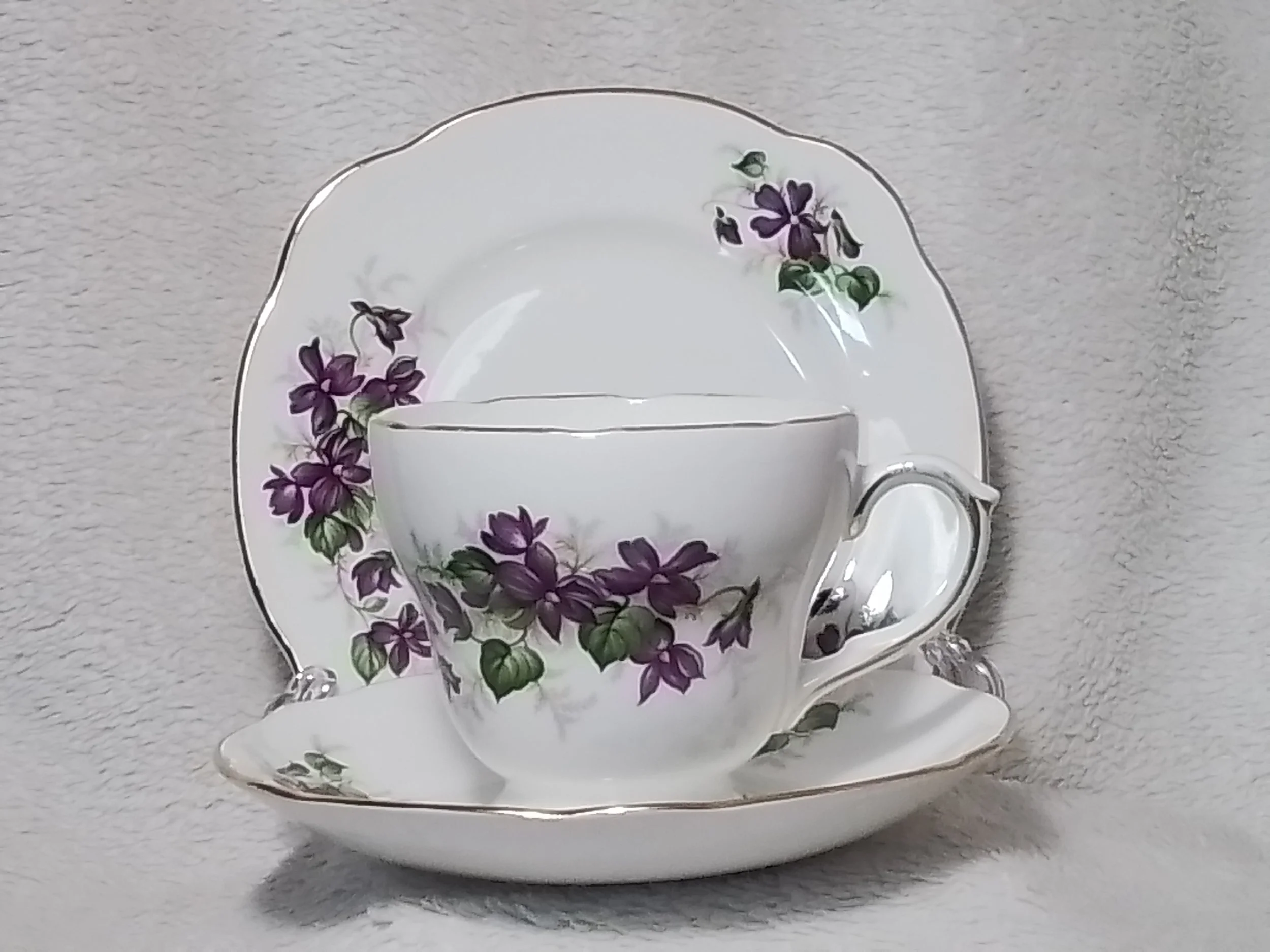 A delicate china teacup and saucer set with purple floral pattern and gold trim, placed on a matching plate, against a neutral background.