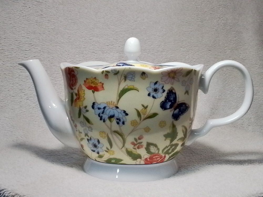 A teapot with a floral and butterfly pattern on a white background, featuring various colored flowers and blue butterflies.