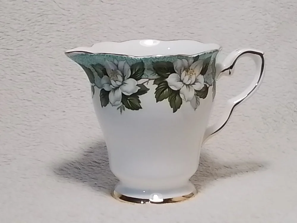 A white porcelain creamer with a floral pattern of white flowers and green leaves, featuring a gold trim, placed against a textured light-colored background.