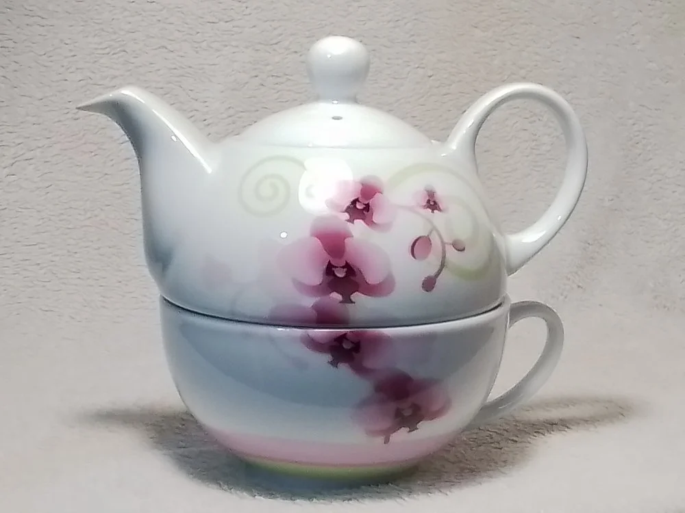 White ceramic teapot with pink and purple floral design stacked on top of a matching teacup with a pink and purple floral design.