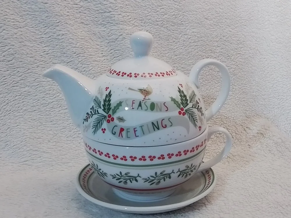 Decorative teapot with holiday design and the words 'Seasons Greetings' on it, sitting on a matching teacup and saucer against a textured beige background.