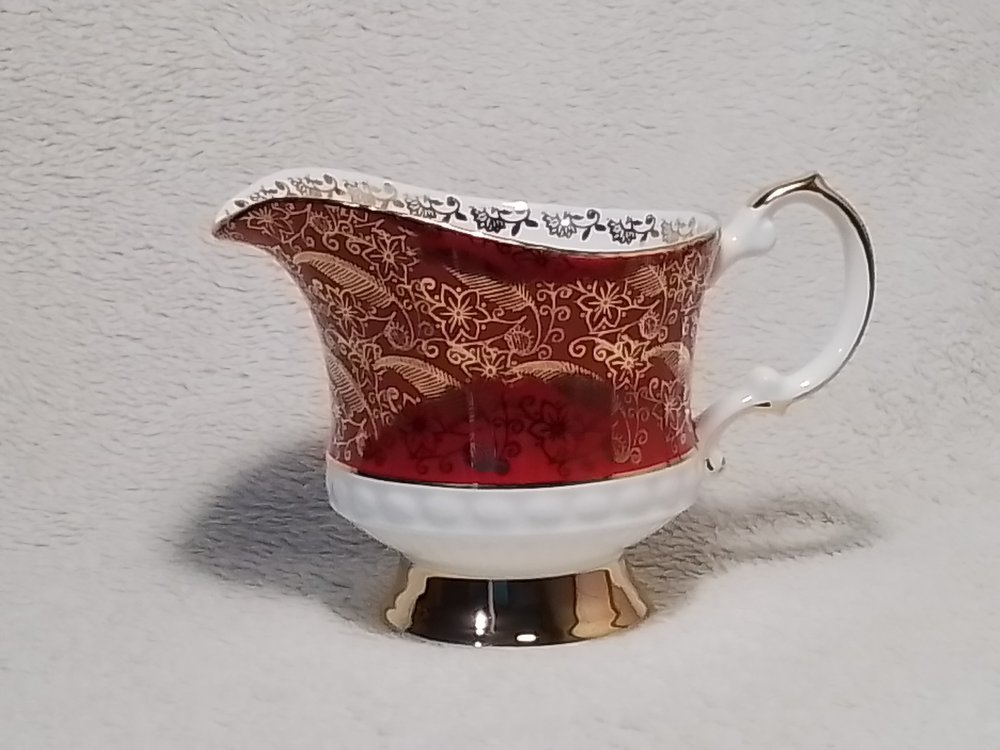 Vintage porcelain gravy boat with red and gold floral pattern on white background, silver trim, and a handle, placed on a light-colored textured surface.