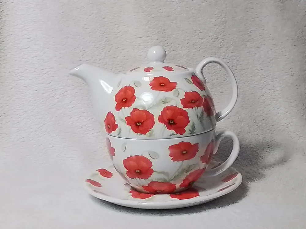 A porcelain teapot and cup set with a matching saucer, decorated with red poppy flower patterns, stacked against a light-colored background.