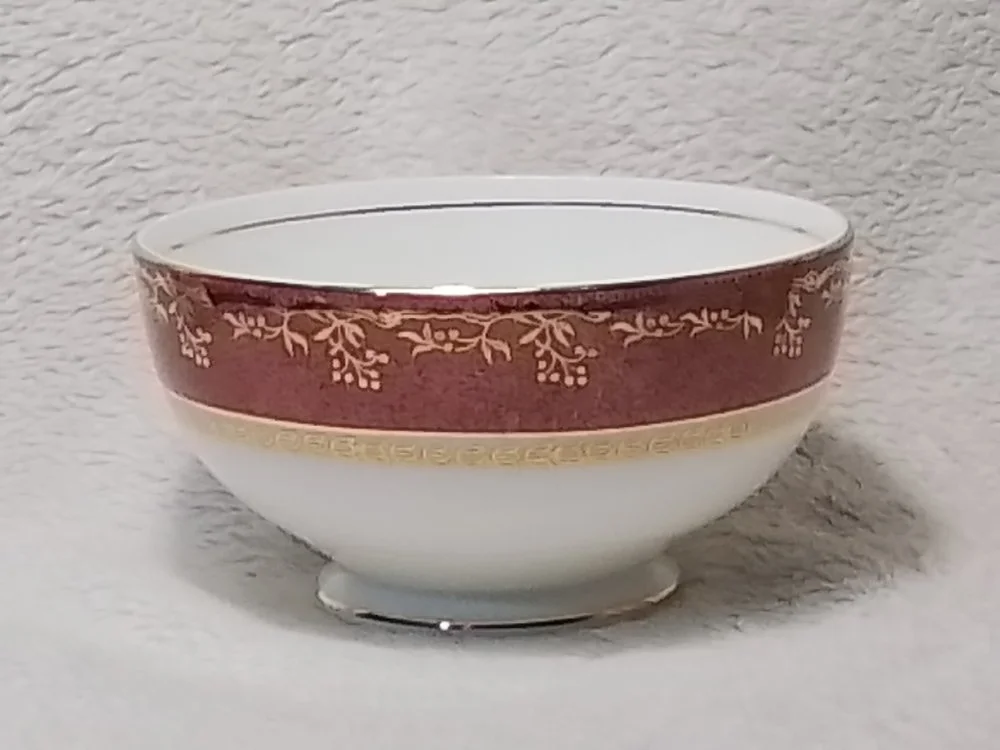 A decorative porcelain bowl with a red and gold floral pattern on the rim and a gold border, placed on a soft, white surface.