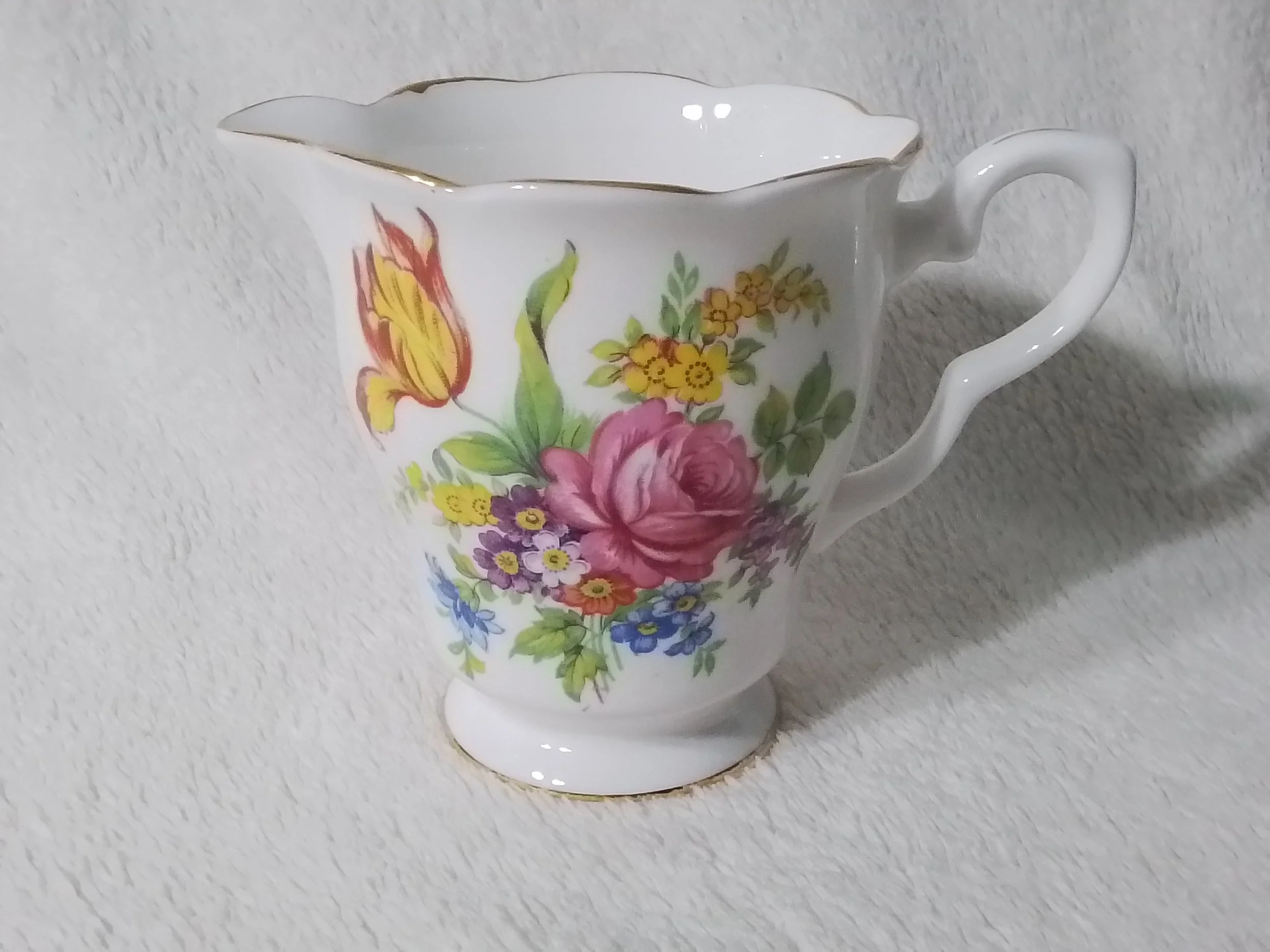 Porcelain creamer with floral pattern and gold trim on handle rim and base, placed on a white textured surface.