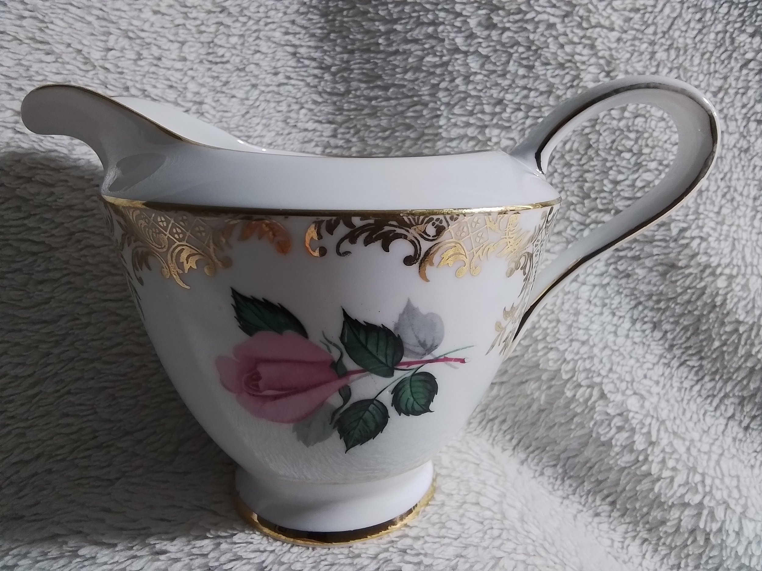 A white porcelain gravy boat with gold trim, decorated with a pink rose and green leaves.