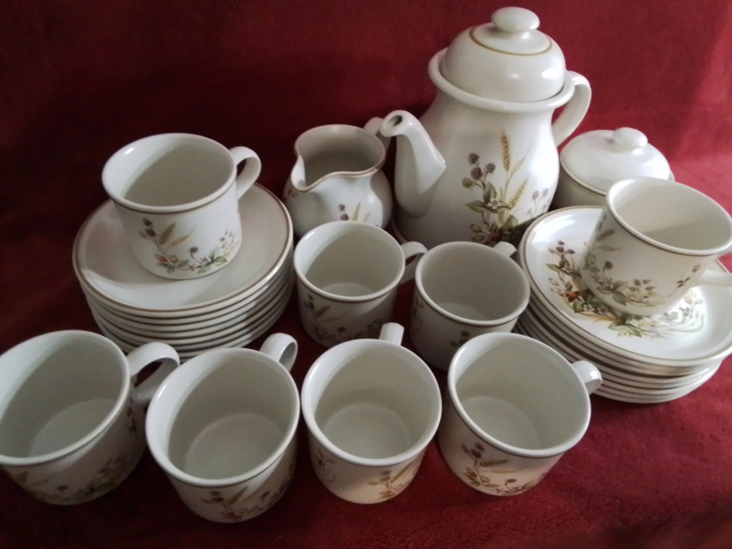 A set of white ceramic teacups, saucers, plates, and a teapot with a floral and leaf pattern, arranged on a red surface.