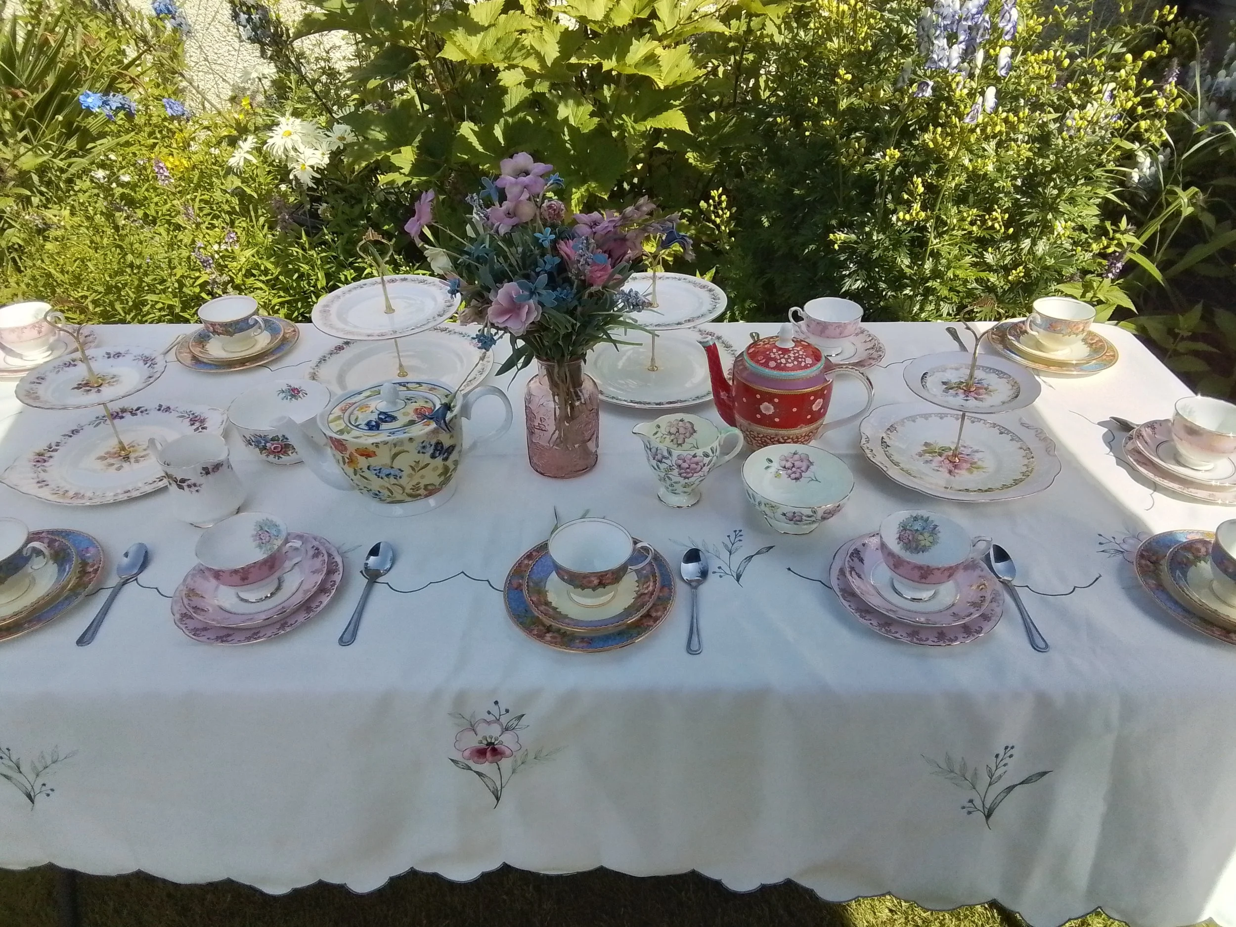 A table set for tea outdoors with floral china, a bouquet of flowers in a pink vase, and greenery in the background.
