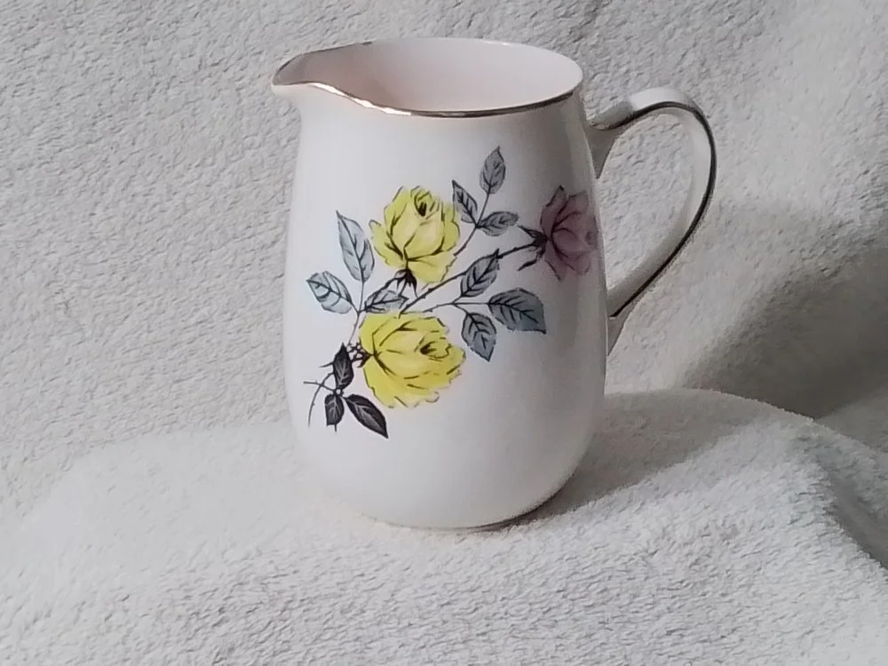 A white ceramic pitcher with a floral design featuring yellow and pink roses with gray and black leaves.