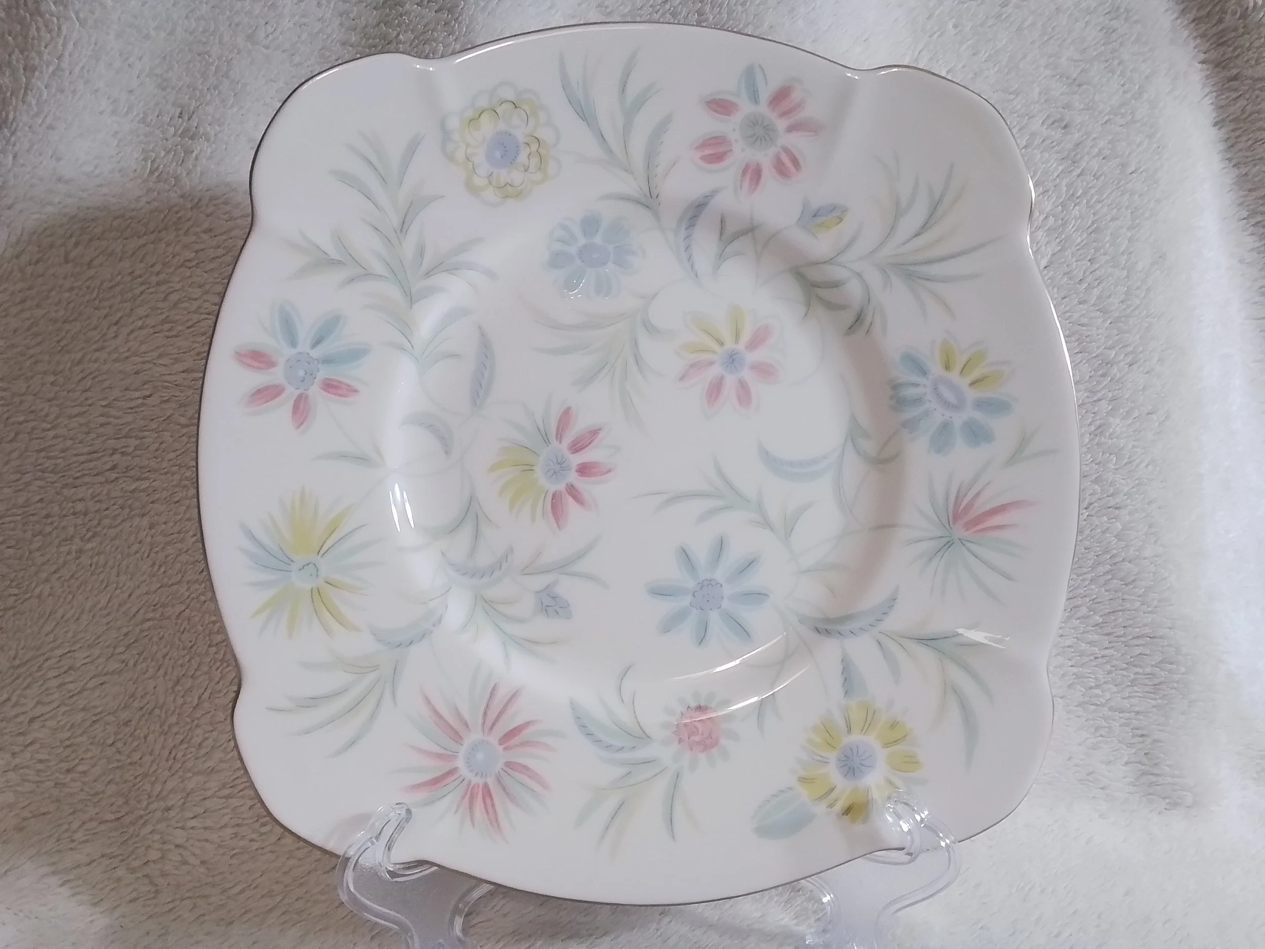A decorative square porcelain plate with a scalloped edge, featuring a pastel floral pattern with pink, blue, yellow, and gray flowers and green leaves, displayed on a clear plastic stand.