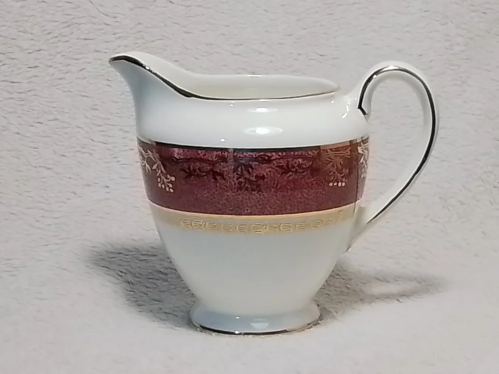 A porcelain creamer with a gold and maroon decorative band around the middle, placed against a textured light background.