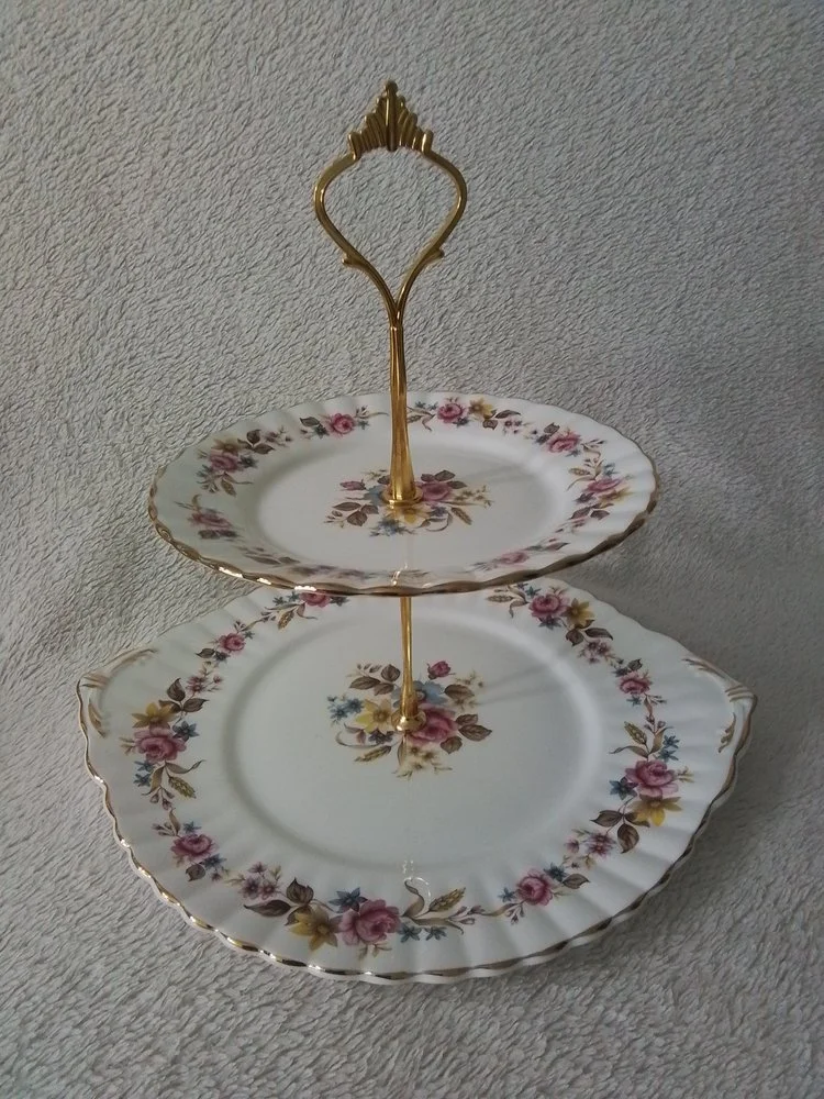 Two-tiered floral and gold-rimmed porcelain serving stand with a decorative handle on top, placed on a textured surface.