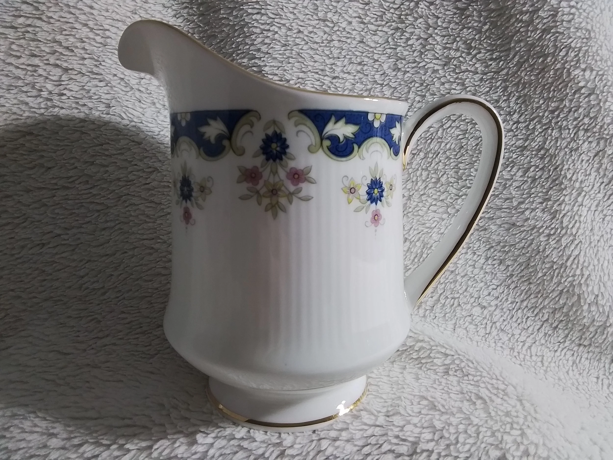 A white ceramic pitcher with floral and gold accents on a textured white fabric background.