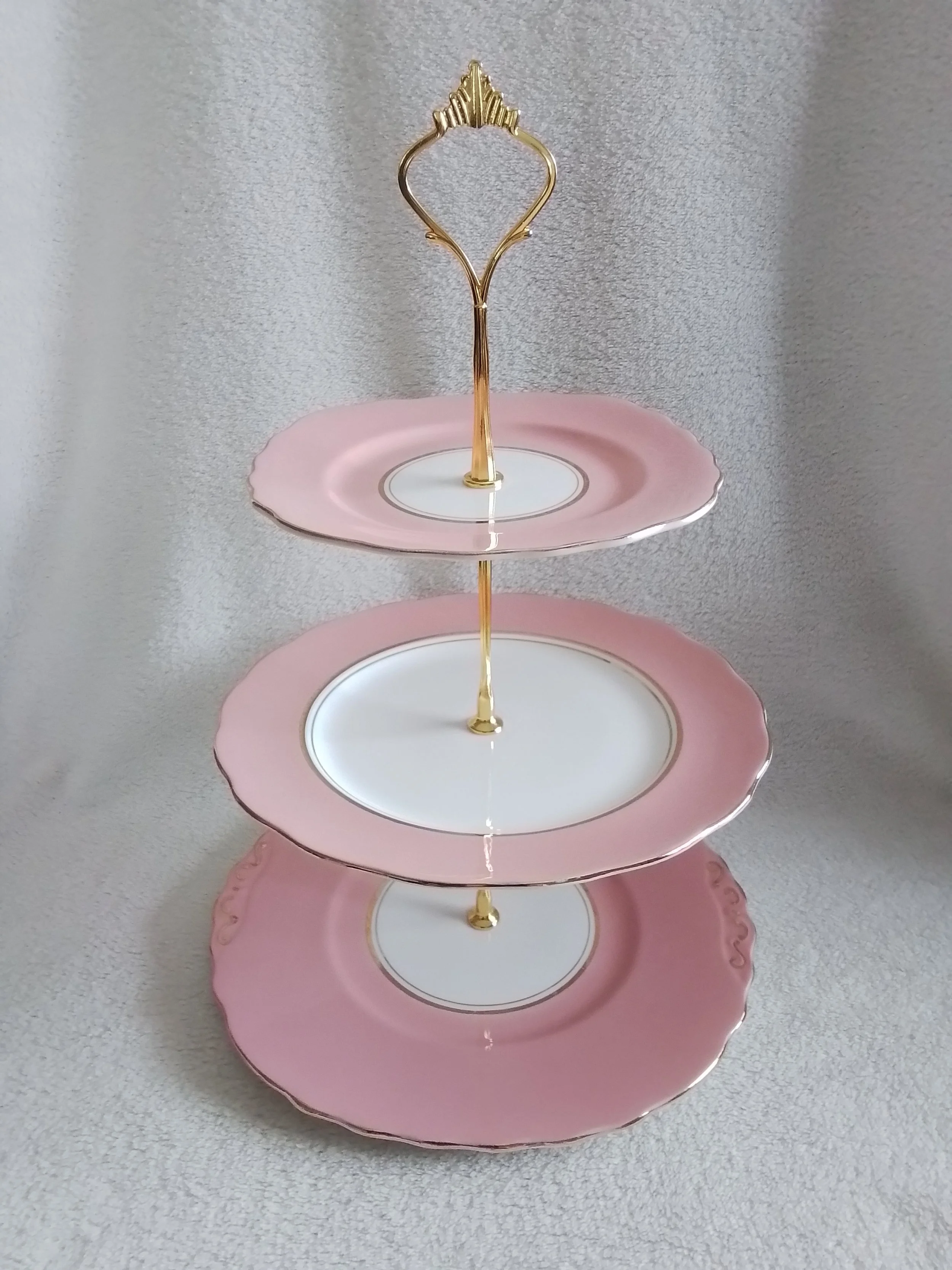 Three-tier pink and white ceramic serving stand with gold accents, empty, against a textured grey background.