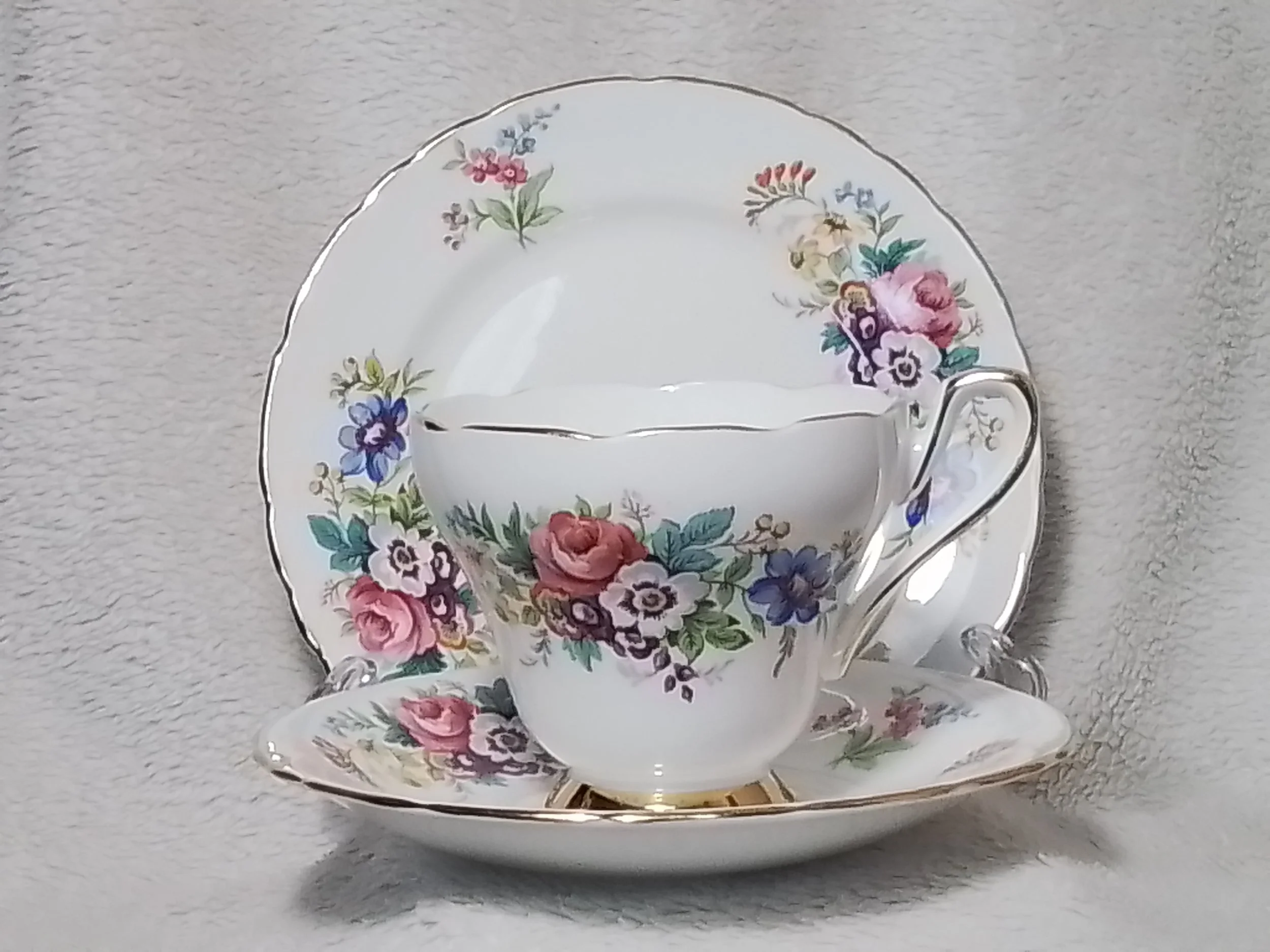 A porcelain tea cup and saucer with floral patterns, featuring pink, blue, purple, and green flowers, set against a light-colored, textured background.