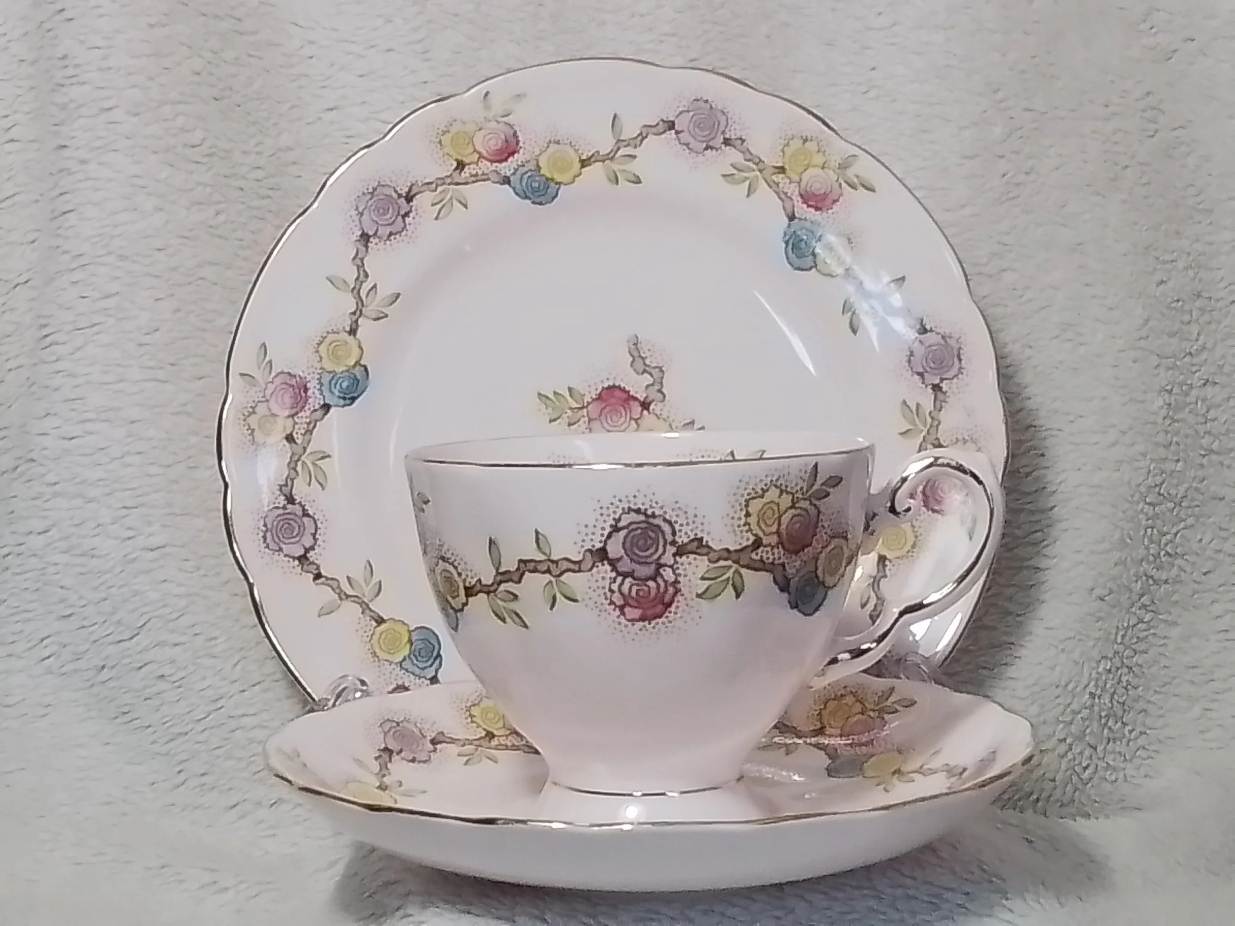 A tea cup, saucer, and dessert plate with a floral pattern featuring pink, yellow, blue, and purple roses and green leaves, set against a light background.