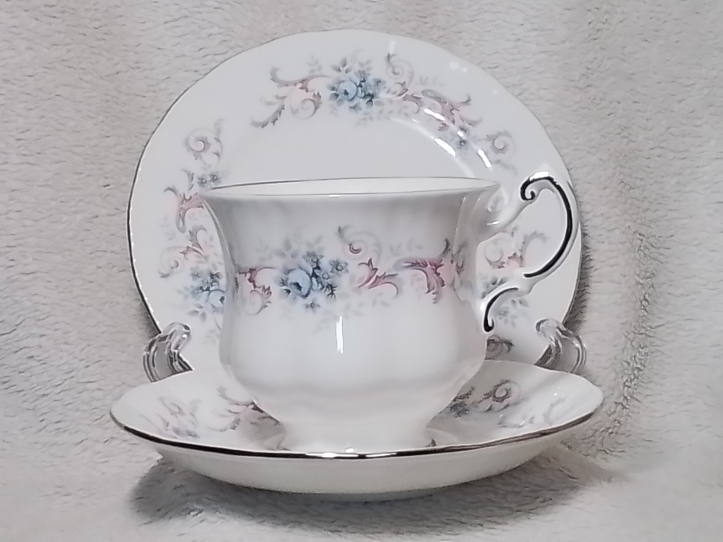 A matching set of porcelain teacup, saucer, and plate with a floral pattern in blue, pink, and gray, with silver trim, displayed against a soft textured background.