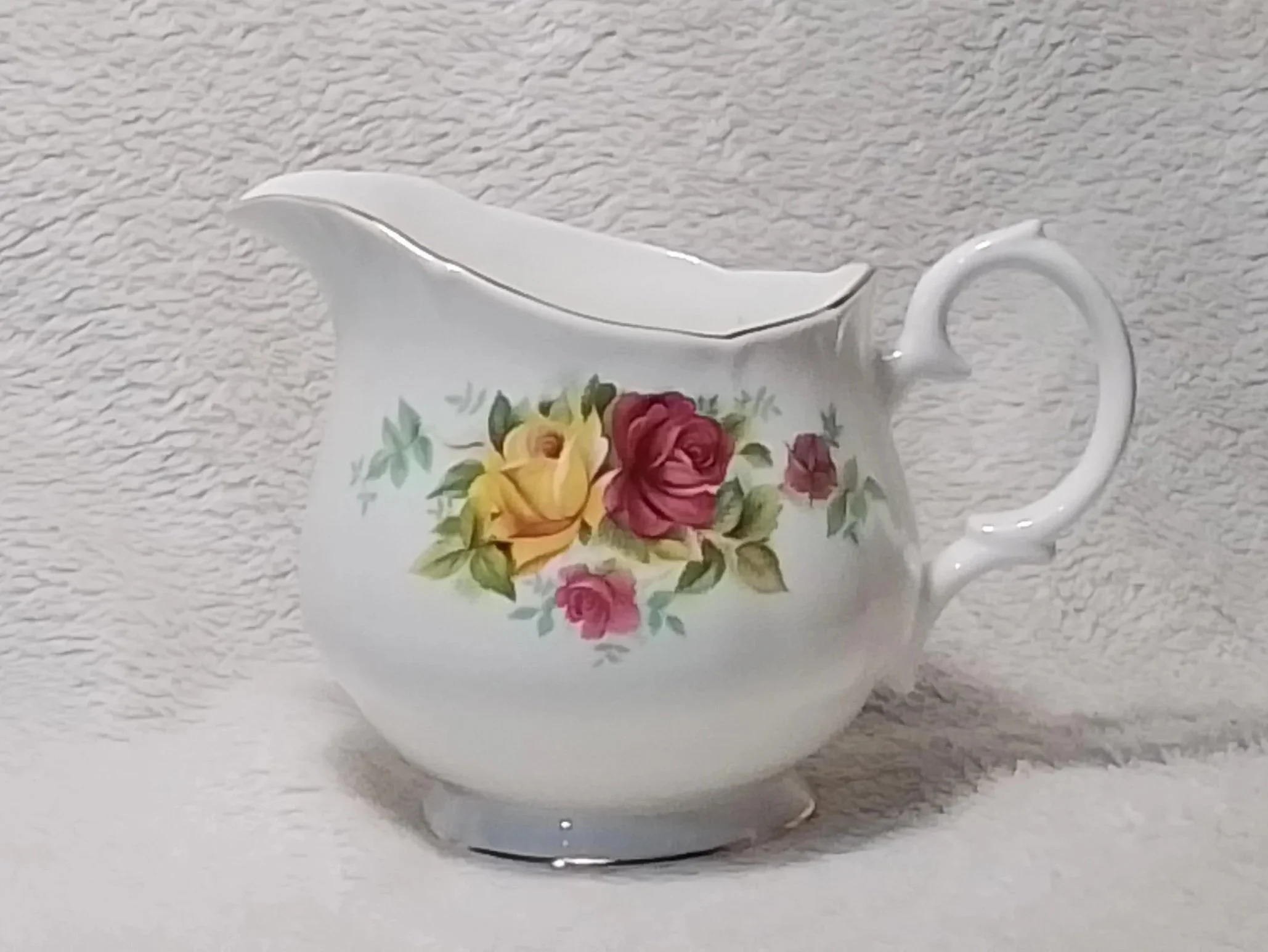 A white porcelain creamer with a floral pattern of yellow, red, and pink roses and green leaves, set against a textured beige background.