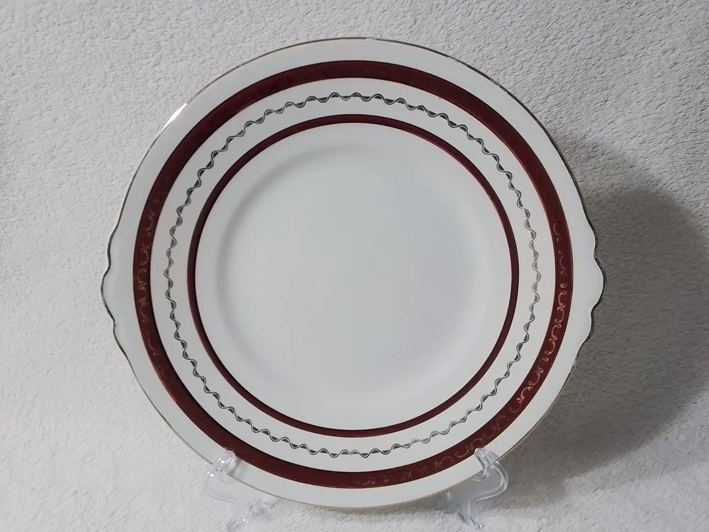 A round ceramic plate with a white center and a decorative red and gold border, displayed on a clear stand against a white textured background.