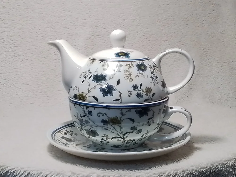 A white ceramic teapot with a floral pattern in blue and green, placed on a matching teacup and saucer set against a light-colored textured background.