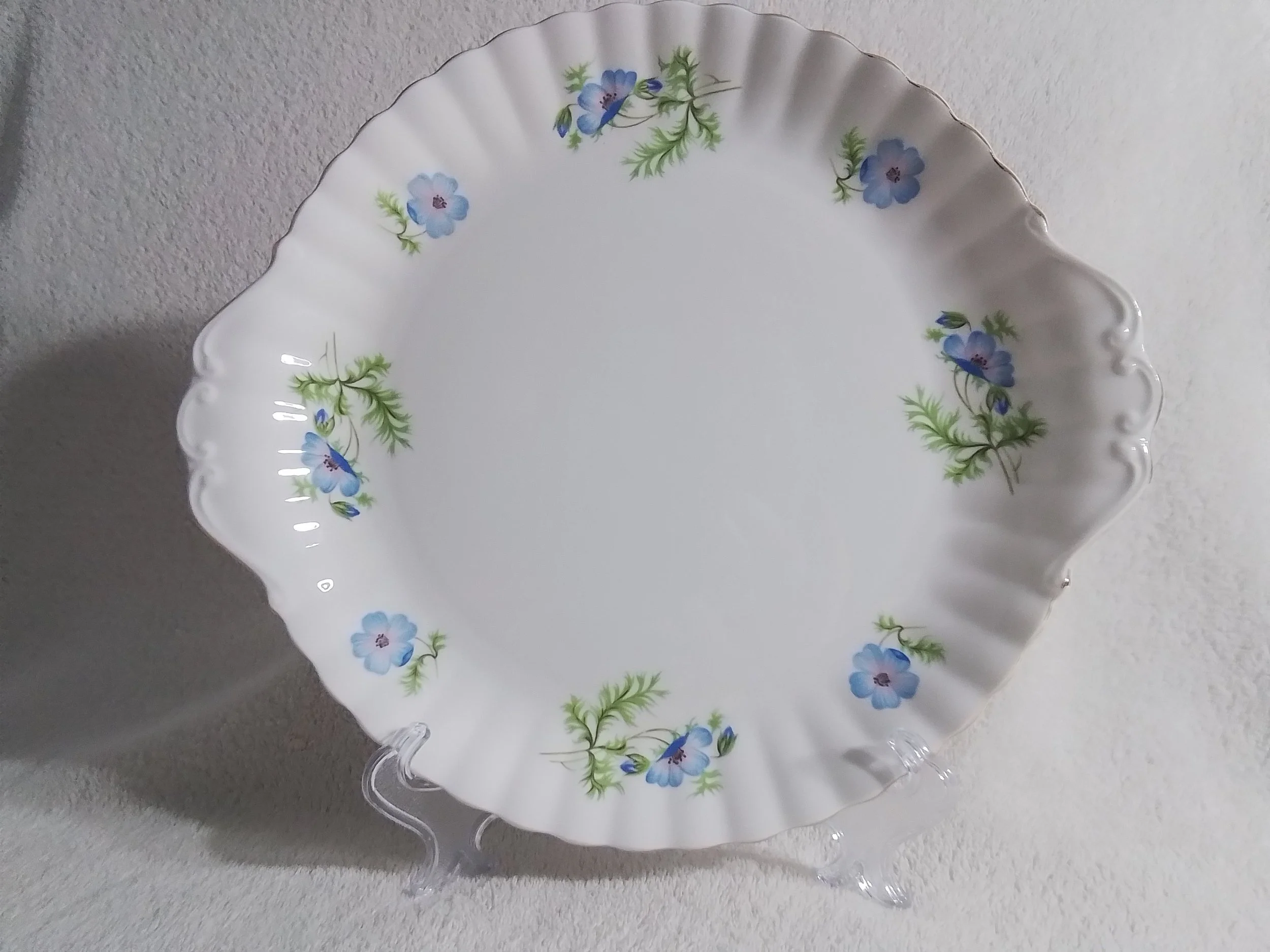 A decorative white porcelain plate with blue flowers and green leaves painted around the rim, displayed on a clear stand against a white background.