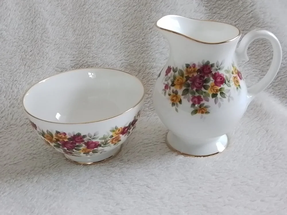 A porcelain bowl and a porcelain creamer pitcher, both decorated with pink, yellow, and purple flowers with green leaves and gold trim, placed on a light-colored textured fabric surface.