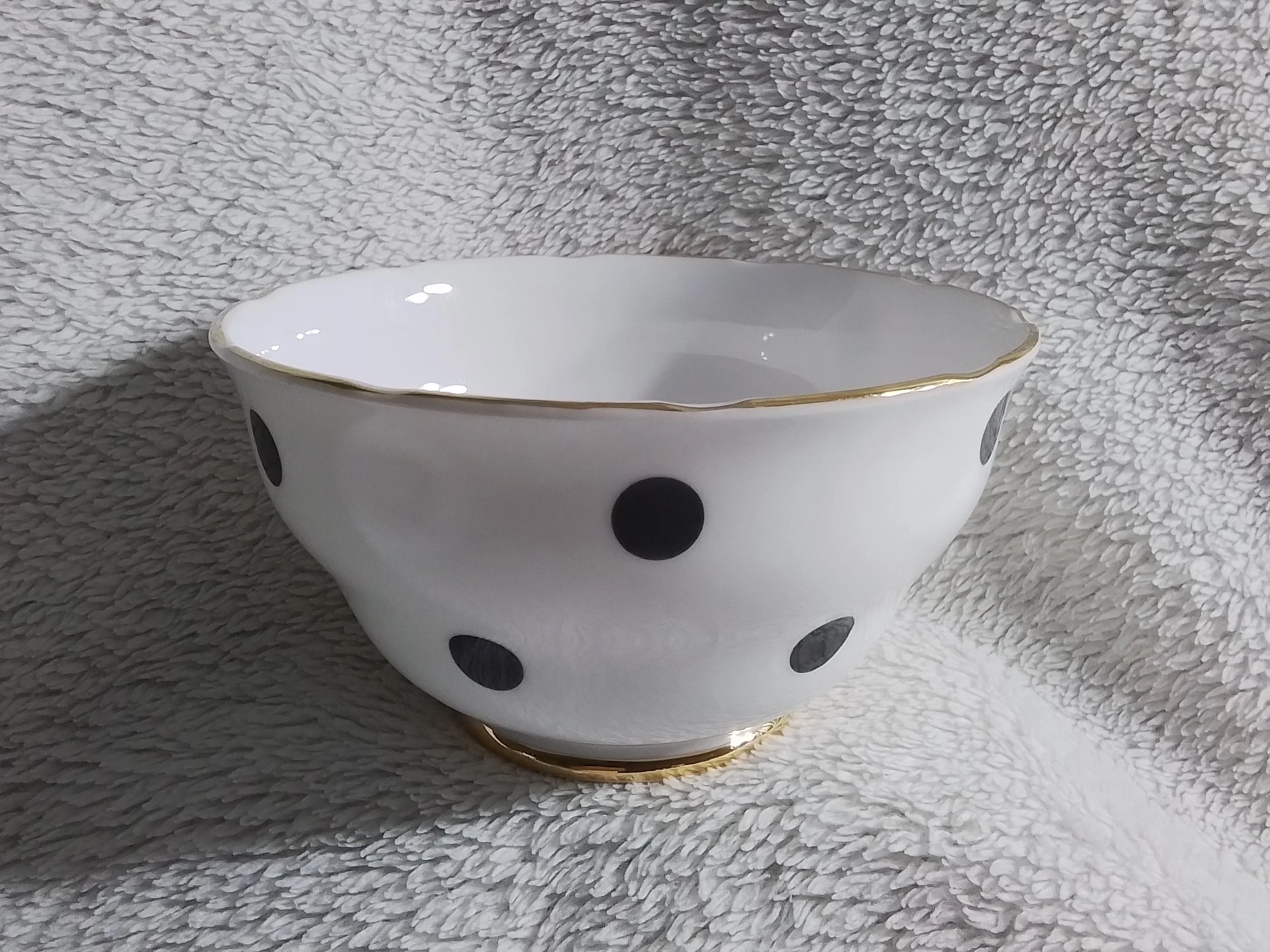 White ceramic bowl with black polka dots and a gold trim, placed on a soft, textured gray fabric surface.