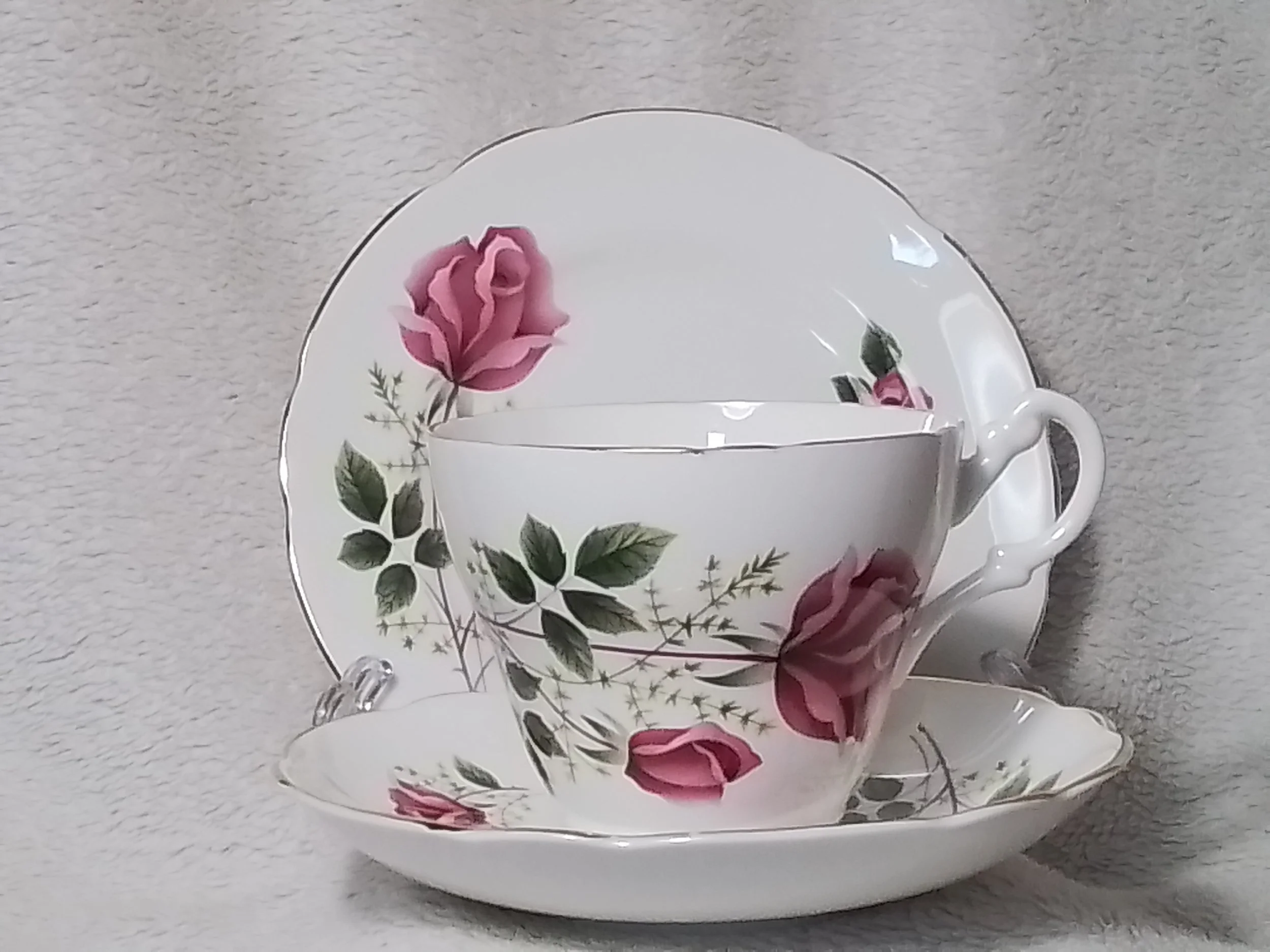 A porcelain tea cup and saucer set decorated with pink roses and green leaves, placed against a light grey textured surface.