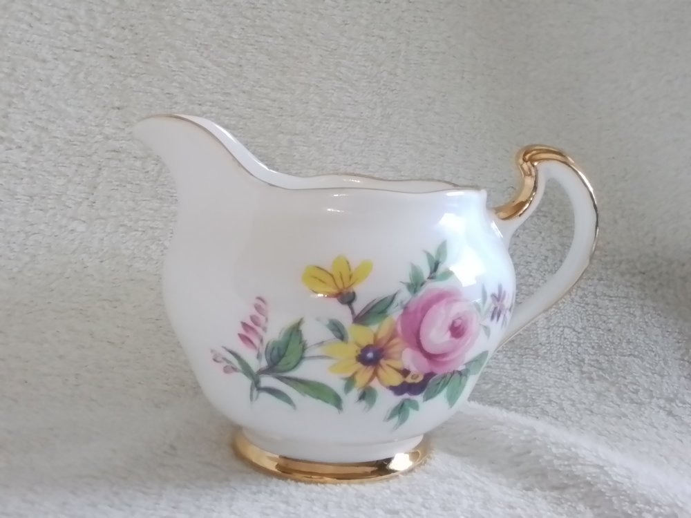 A white ceramic creamer pitcher with floral pattern and gold accents on the handle and base, placed on a textured surface with a neutral background.
