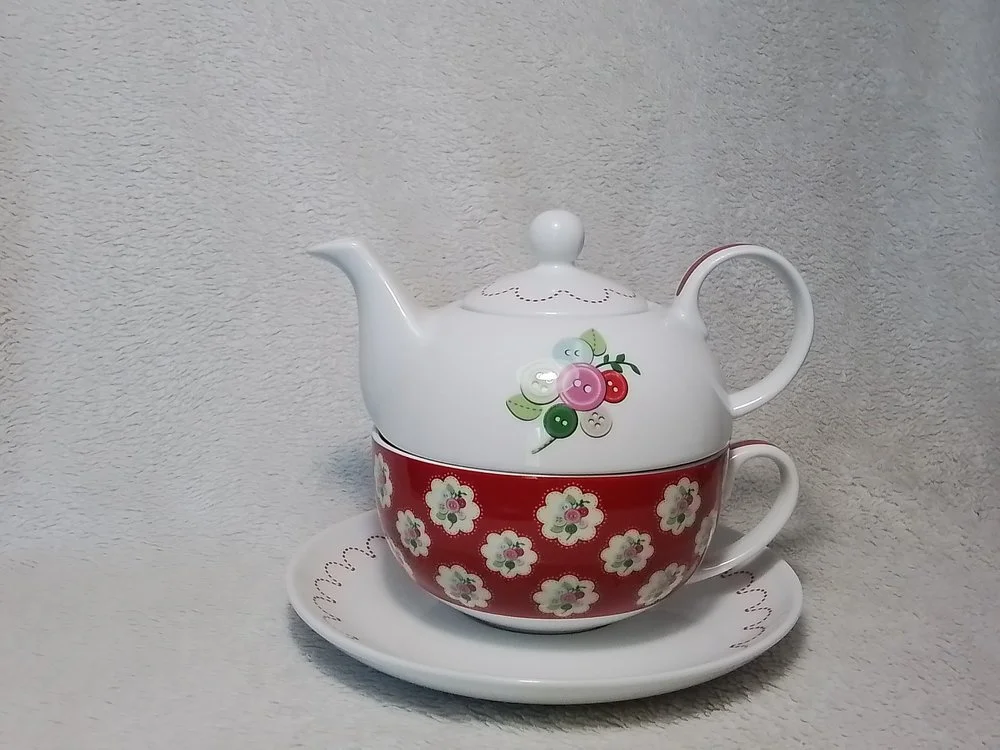 A decorative teapot and cup set with floral patterns, placed on a matching saucer. The teapot has a white top with a flower and berry appliqué, and a red band with white flowers around the middle.