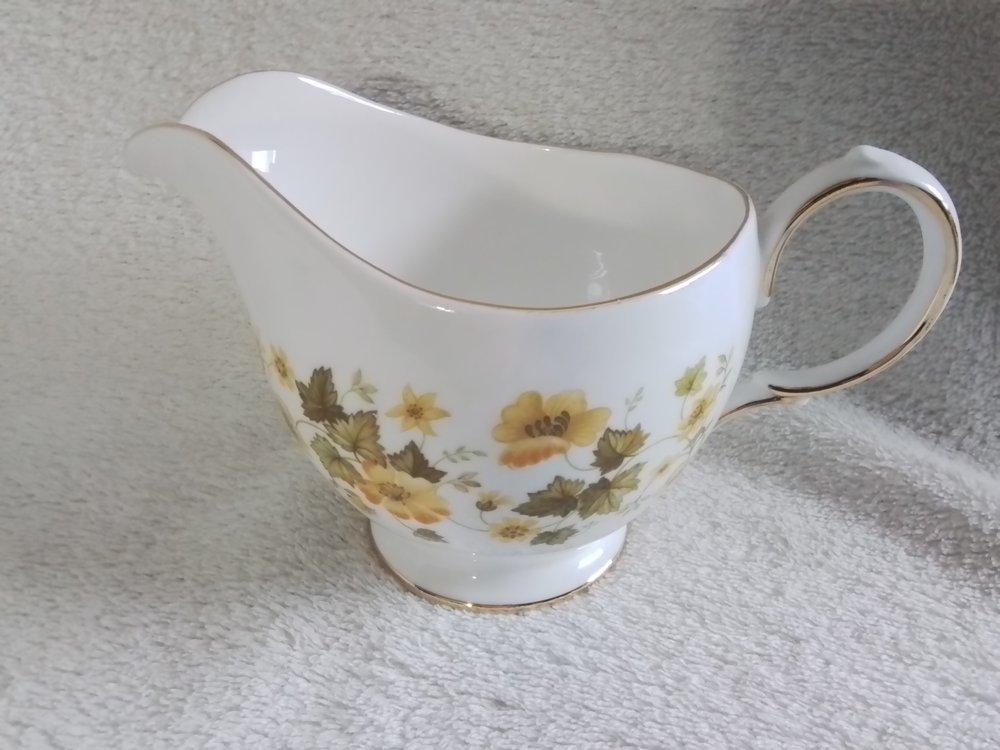 A white porcelain gravy boat with gold trim and floral pattern of yellow and brown flowers and green leaves, placed on a light-colored textured surface.