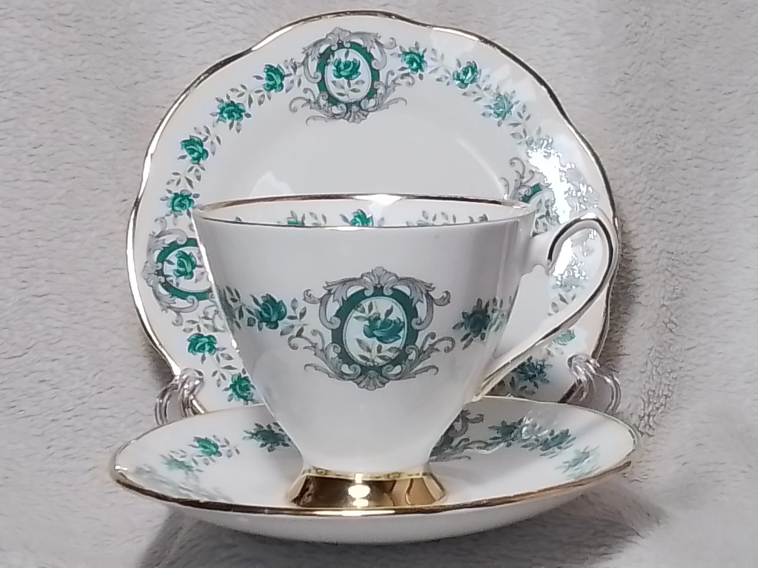 A fine china tea set with a white background and teal and gray floral patterns, including a teacup, saucer, and a dessert plate with gold trim.