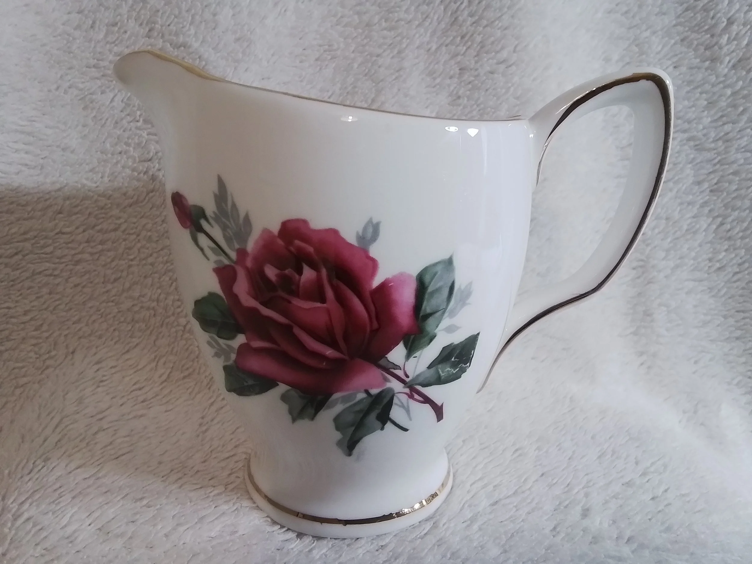A white ceramic vase with a pink rose floral design and gold accents on the rim, handle, and base, placed on a soft, textured white surface.