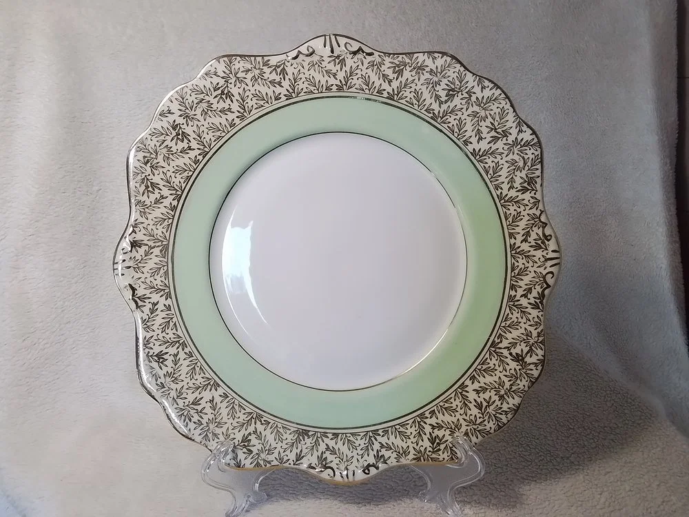 A decorative porcelain plate with a floral black and white pattern on a scalloped edge, featuring a light green band near the rim, displayed on a clear plastic stand.