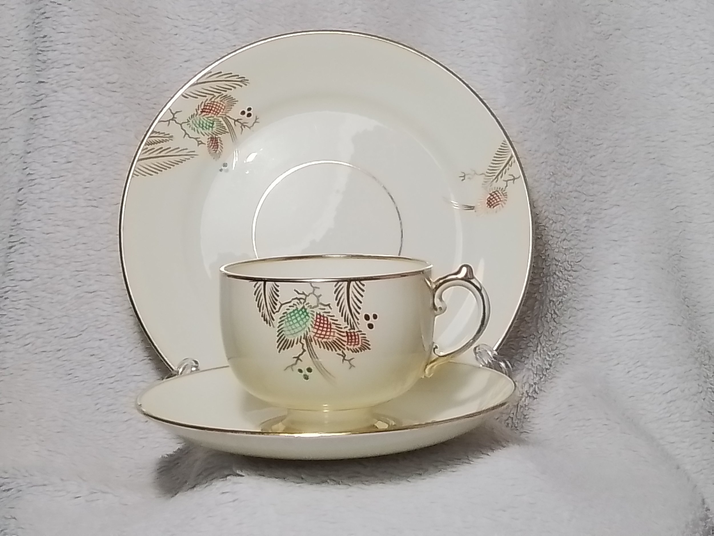 A cream-colored china tea cup, saucer, and large plate with a holly berry and leaf pattern with gold accents, arranged on a textured gray background.
