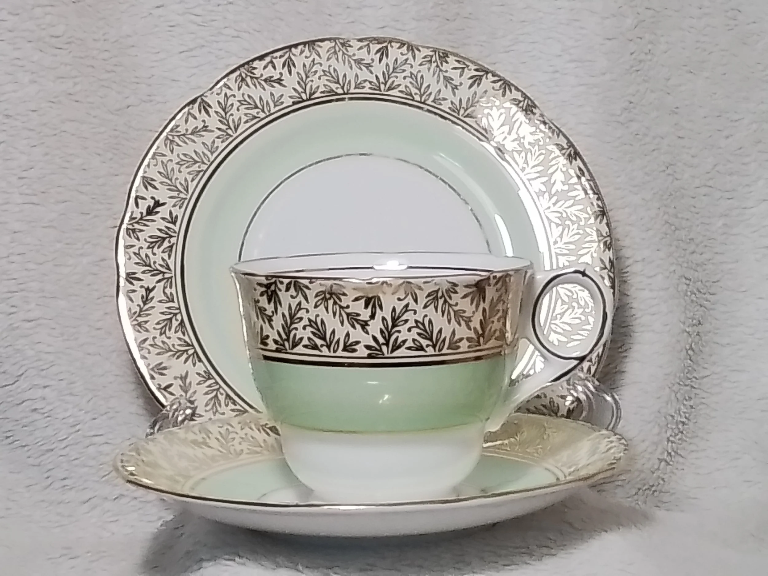 Elegant tea set with a cup, saucer, and a large plate, all featuring matching black and white leaf patterns and gold accents, displayed against a soft gray textured background.