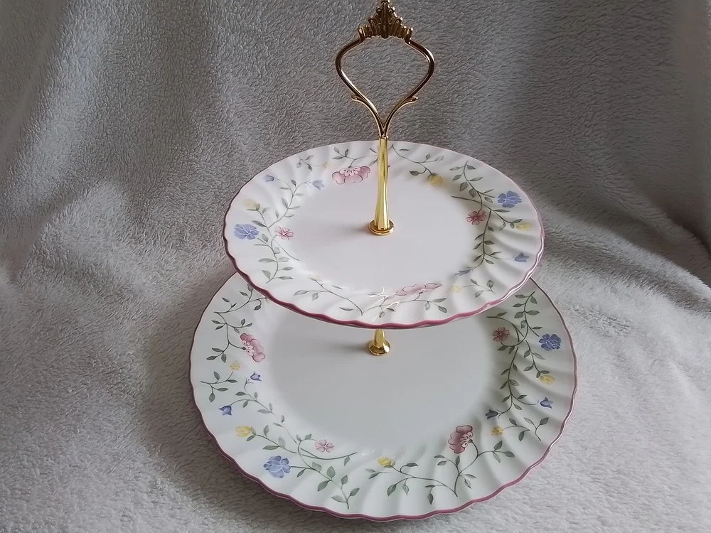 Two-tiered porcelain serving tray with floral pattern, gold accents, and a decorative handle, placed on a soft, light-colored fabric background.