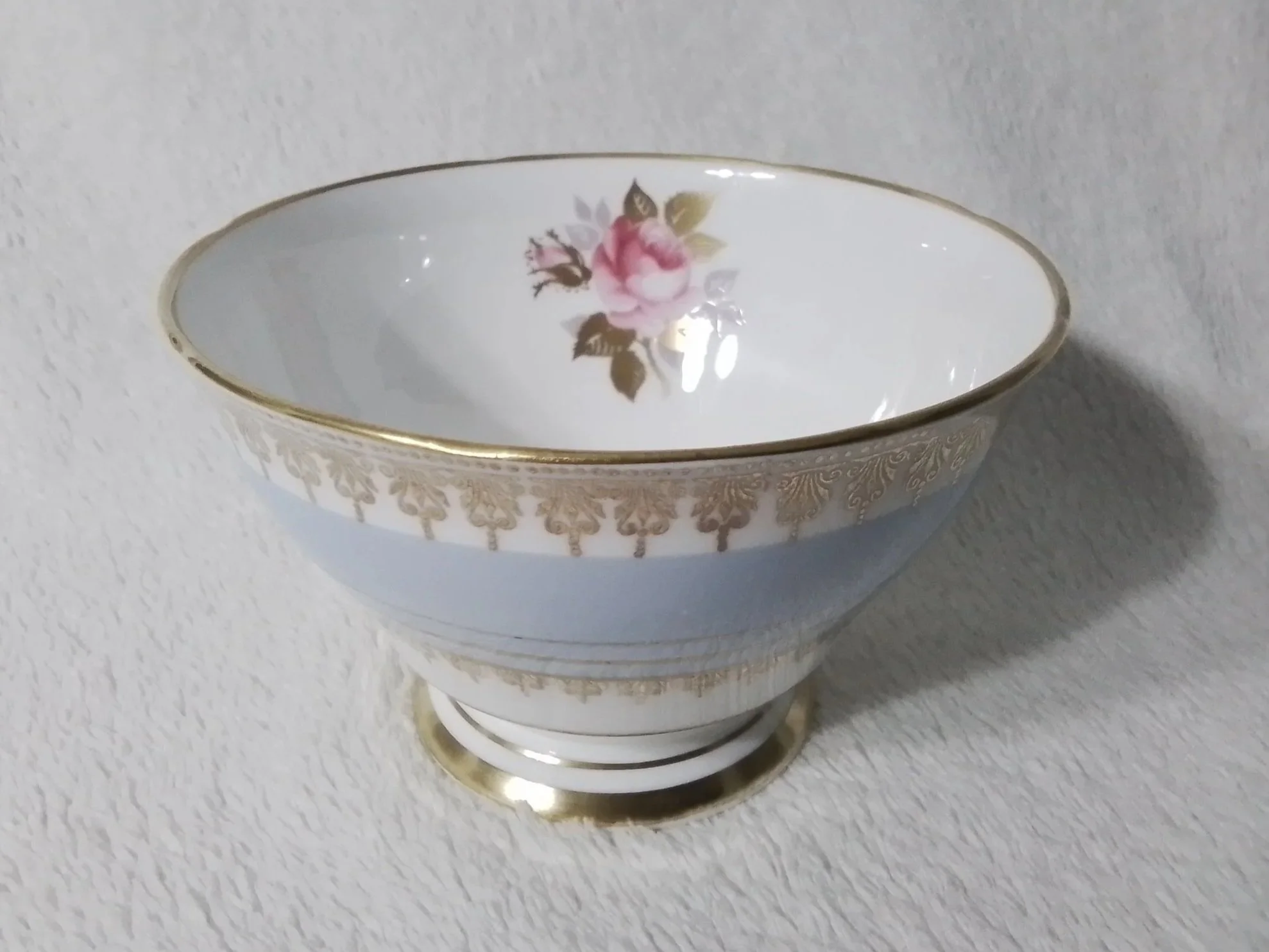 A decorative porcelain bowl with a pink rose and green leaves painted inside, gold trim, and a gold pattern around the exterior, placed on a white textured surface.