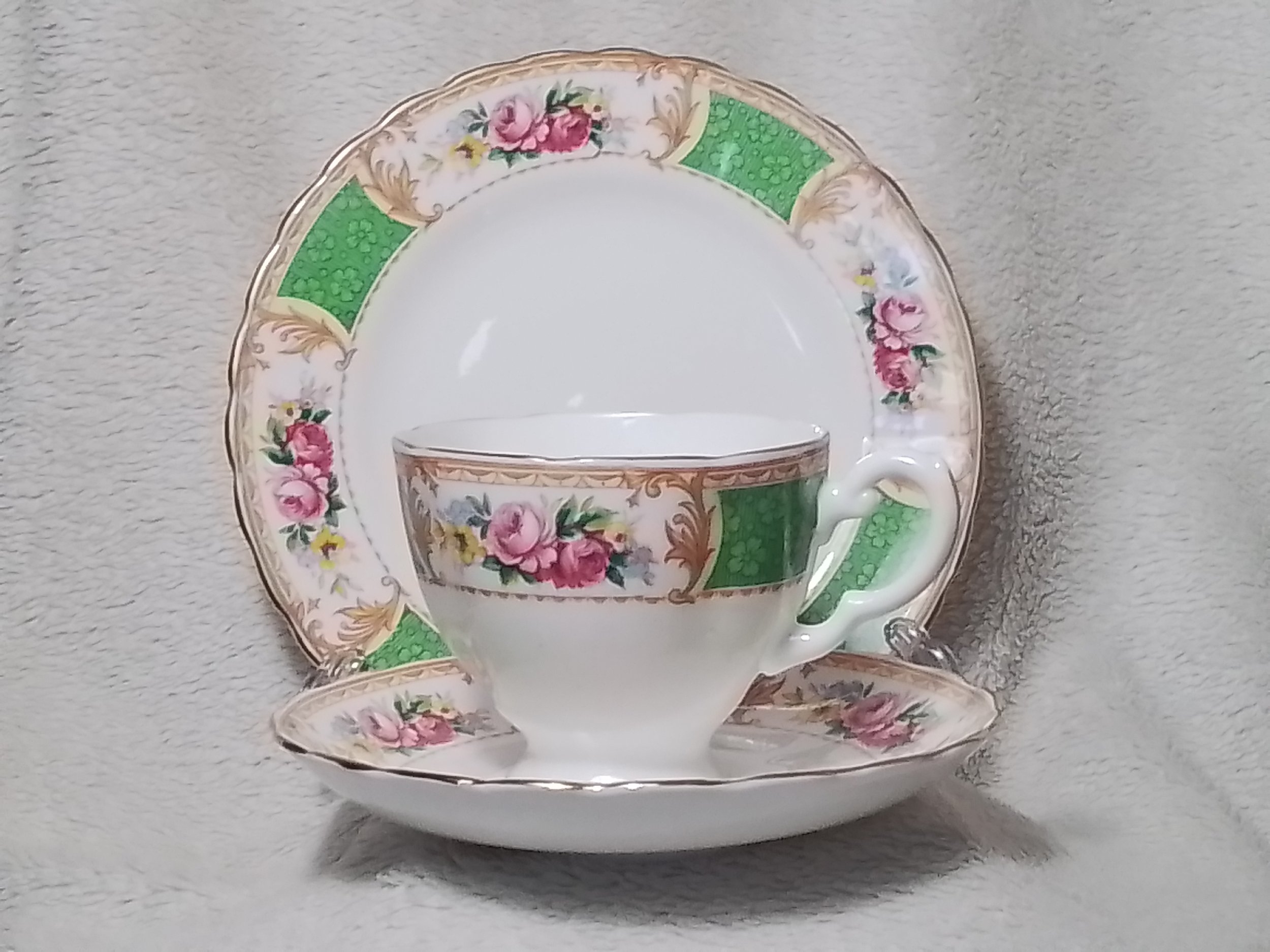 Elegant vintage china teacup, saucer, and dinner plate set with floral pink roses, green accents, and gold trim, displayed on a light textured surface.