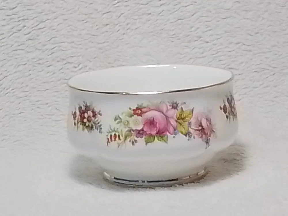 A white porcelain bowl with a floral pattern of pink and purple flowers, and green leaves, and a silver rim around the top edge, placed on a plain white surface with a textured white wall background.