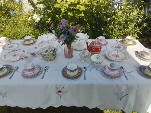 A table set for a meal with floral teapot, cups, and saucers, white plates, and silverware outdoors amid greenery and flowers.