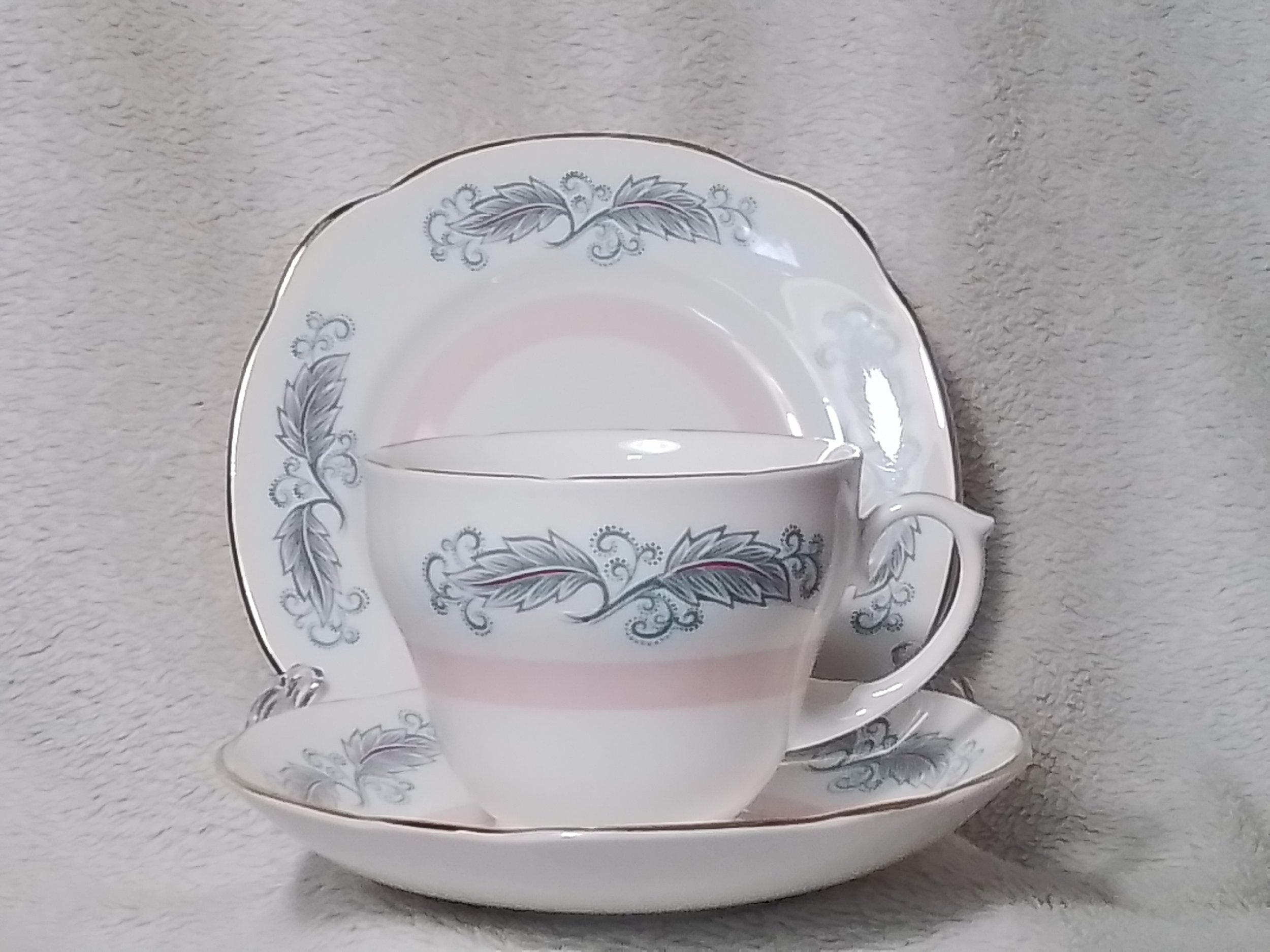 A fine china teacup with a matching saucer and a decorative plate, all with a floral pattern in gray and pink on a white background.