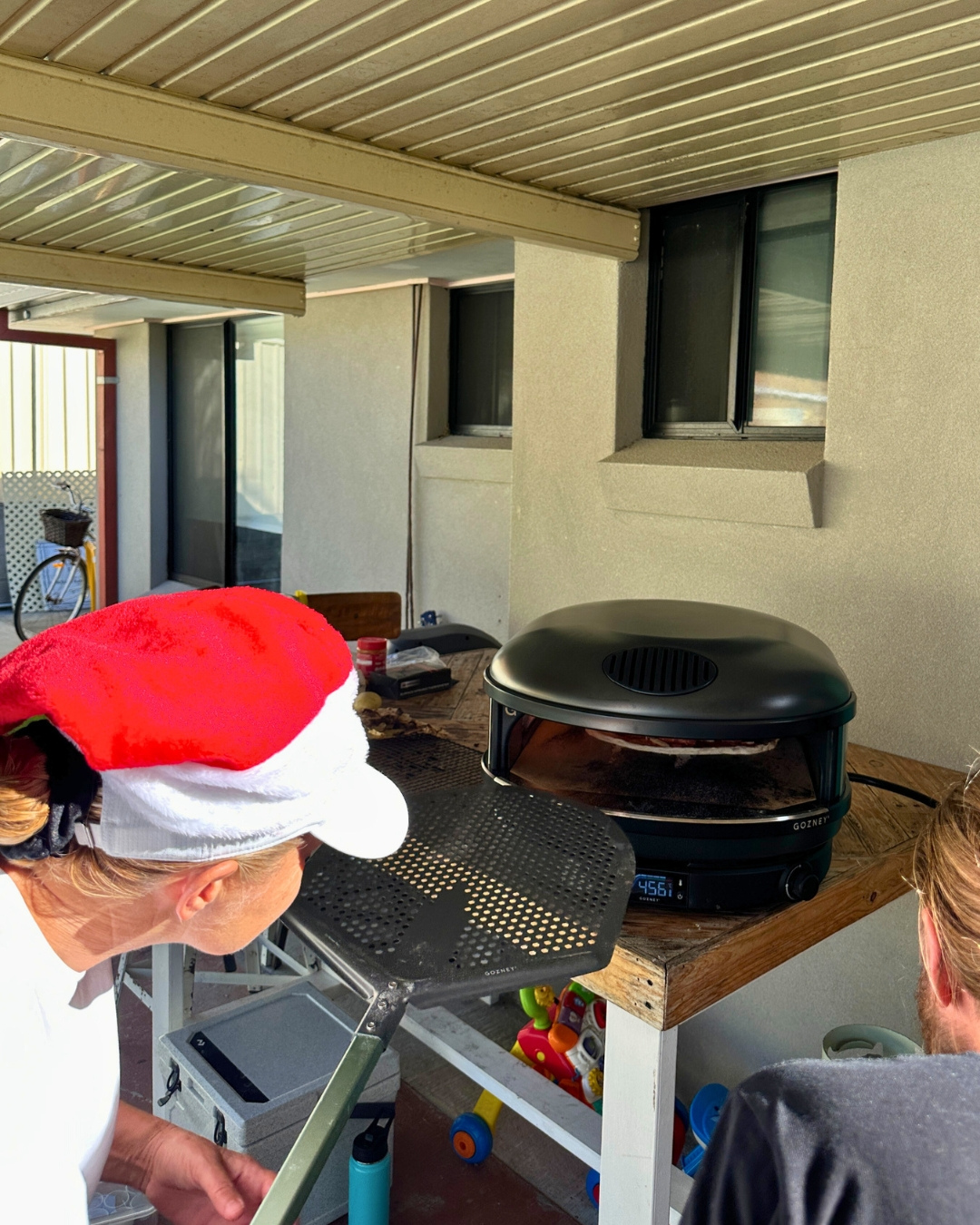 A guest in a Santa hat peers into a Gozney pizza oven during a Marino & The Dough masterclass, as a pizza cooks on the stone. A relaxed backyard setting shows hands-on learning and festive fun.